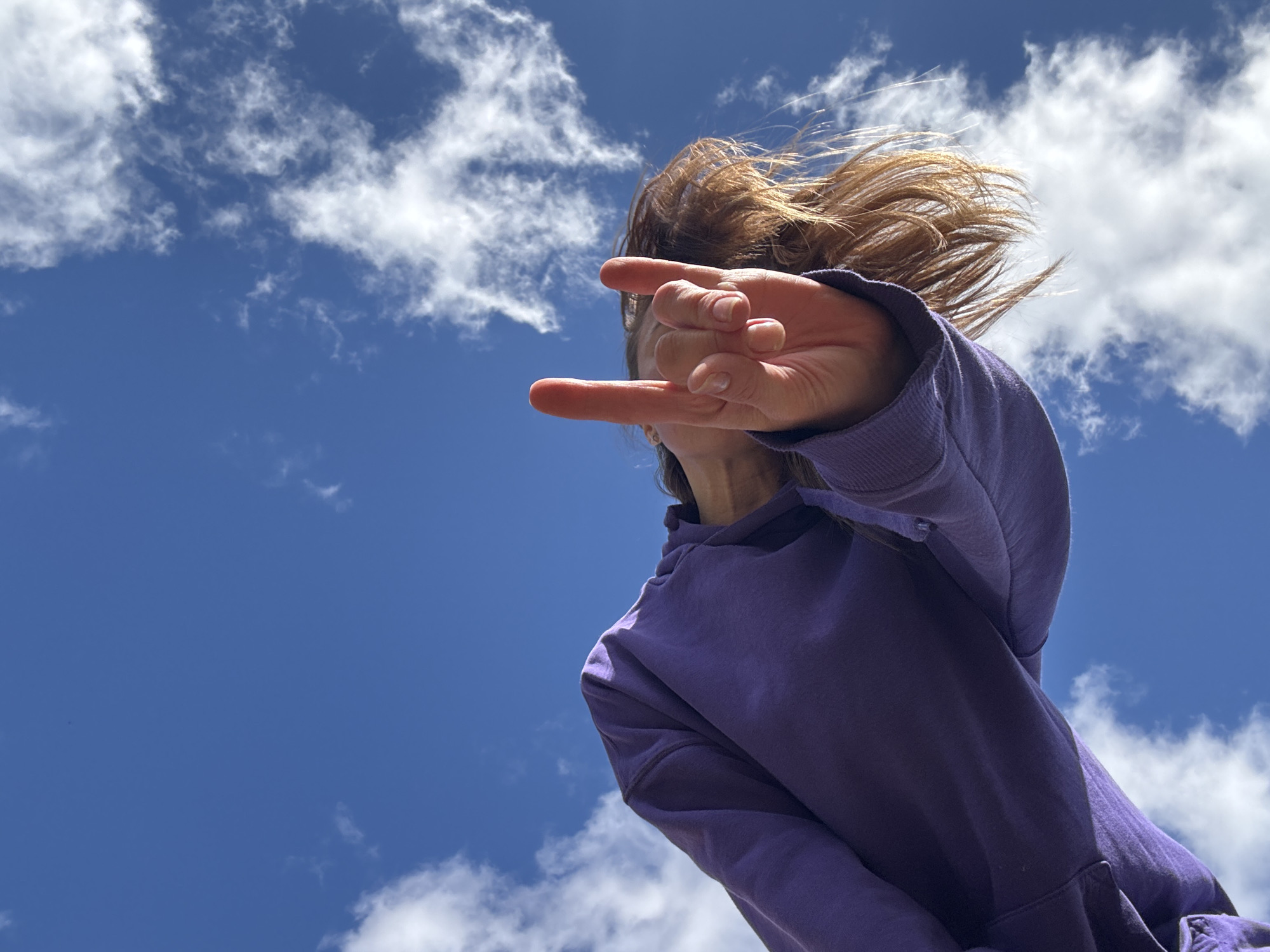 The image shows a person, likely female, wearing a purple hooded sweatshirt, viewed from a low angle against a backdrop of a bright blue sky scattered with white cumulus clouds. The person's face is partially obscured by their hand, which is held up in a gesture where the index and pinky fingers are extended, resembling the "rock on" or "devil horns" hand sign. The subject's hair is light brown and appears to be blowing slightly in the wind. The environment appears to be outdoors on a sunny day. There is no evidence to suggest that the location is in the city of Прикамский, Russia.
