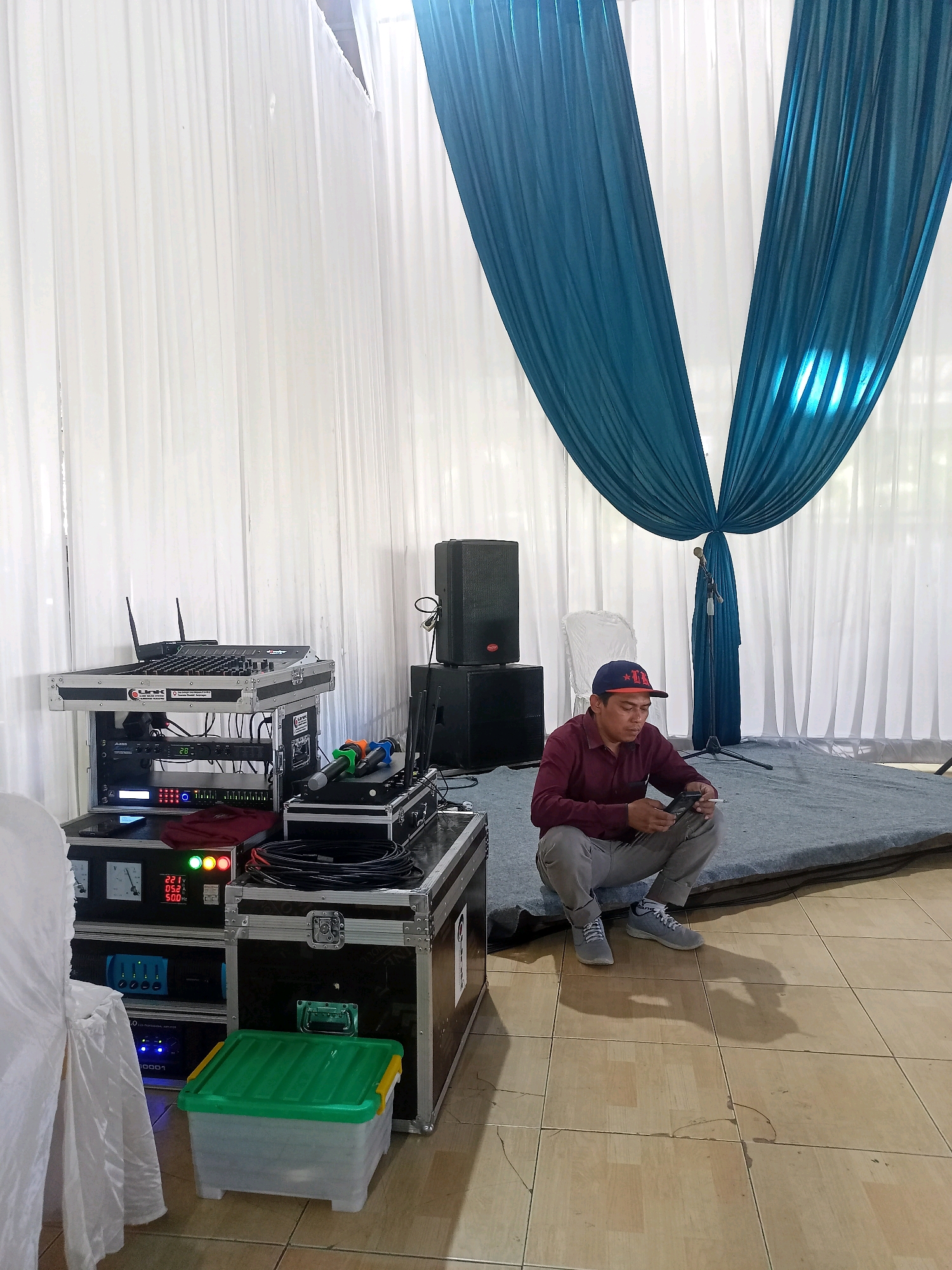 A man wearing a maroon shirt, grey pants, a baseball cap, and grey sneakers is squatting on a grey carpeted floor, looking at his mobile phone. He is holding a cigarette in his right hand. Behind him, white curtains are draped, with teal fabric hanging in front of them. To his left, a stack of audio equipment, including speakers and a mixing console, is set up. A white chair is visible behind the man. The floor is tiled. The scene appears to be a venue or event space in Wanadadi, Indonesia, likely set up for a performance or gathering.