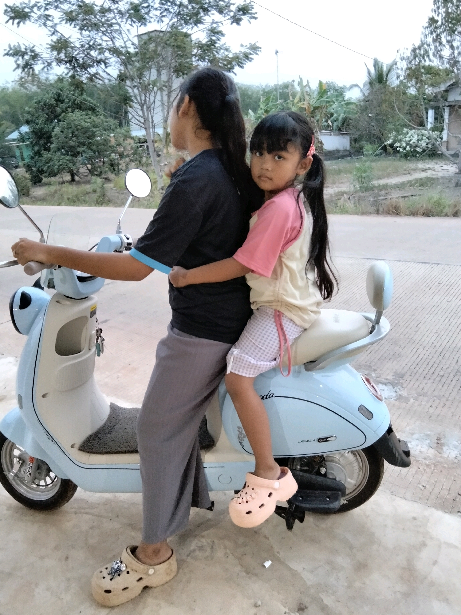 In Perambahan, Indonesia, two individuals, an adult and a child, are positioned on a light blue scooter. The adult, wearing a dark t-shirt with blue trim and gray pants, is seated in front, holding the handlebars. The child, in a pink and white shirt and pink checkered shorts, sits behind the adult, with their arms around the adult's waist. Both are wearing clogs. The scooter is parked on a concrete surface, with a natural background of trees and some buildings visible. The overall lighting suggests it is either early morning or late afternoon.