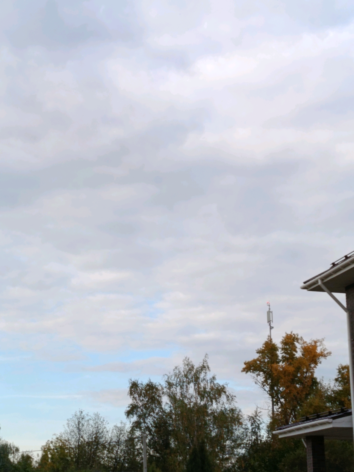 The image shows a view of the sky with clouds, trees, and part of a building. In the background, a cell tower with a red light at the top is visible among the trees. The building in the foreground appears to be a house with a white soffit and gutter system. The trees show signs of autumn foliage, with some leaves displaying yellow and orange colors. The lighting suggests it is daytime. There are no people or animals visible in the image. The location is identified as Vereya, Russia.