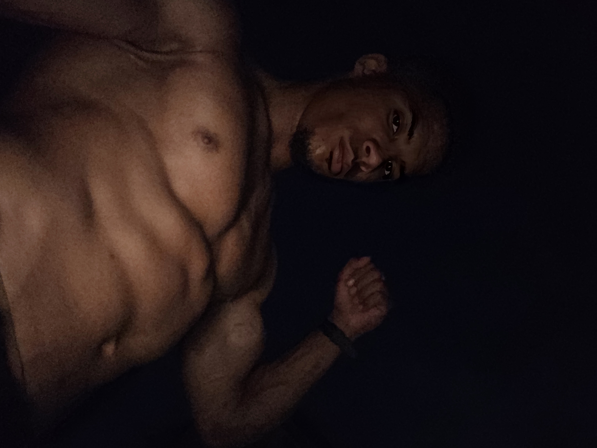 The image shows a low-angle, close-up shot of a muscular, shirtless man against a dark background. The man has dark skin, short dark hair, and is looking towards the camera with a serious expression. His right arm is flexed, and he is wearing a dark wristband. The lighting highlights his abdominal muscles and chest. The environment is dark and lacks distinguishing features, making it impossible to determine a specific location within Varna, Bulgaria.