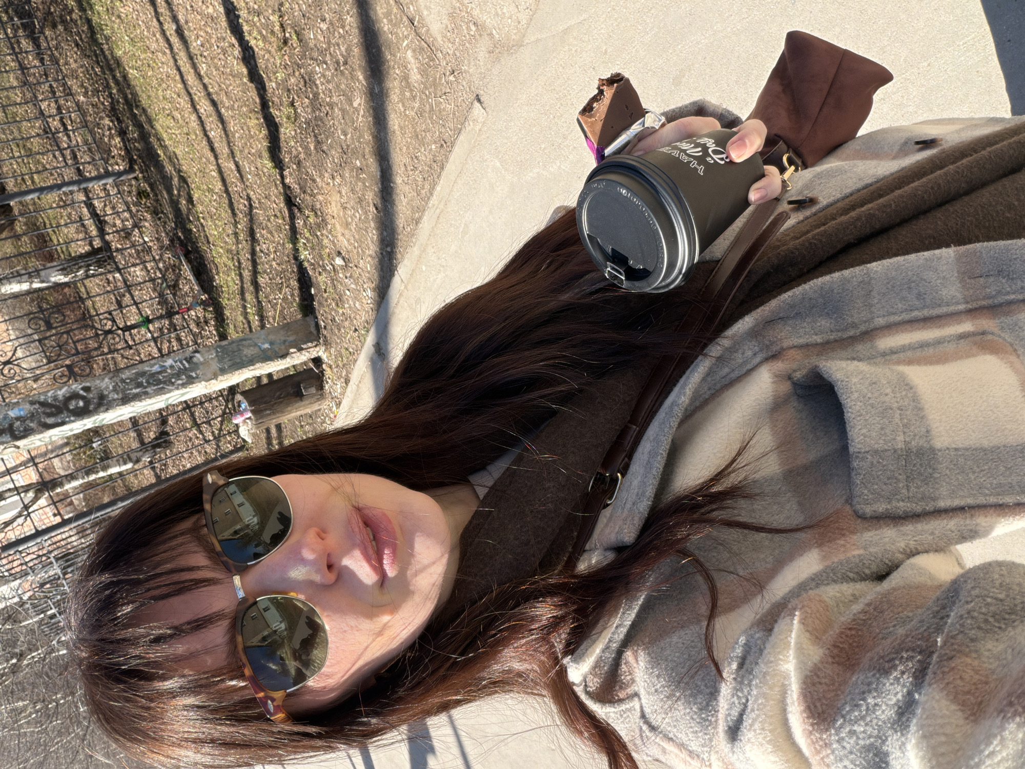 A selfie captures a woman with long brown hair, wearing sunglasses and a brown, plaid jacket, holding a coffee cup and a piece of chocolate. The background shows a fence and foliage. This is likely a selfie taken by the woman in Perm, Russia.