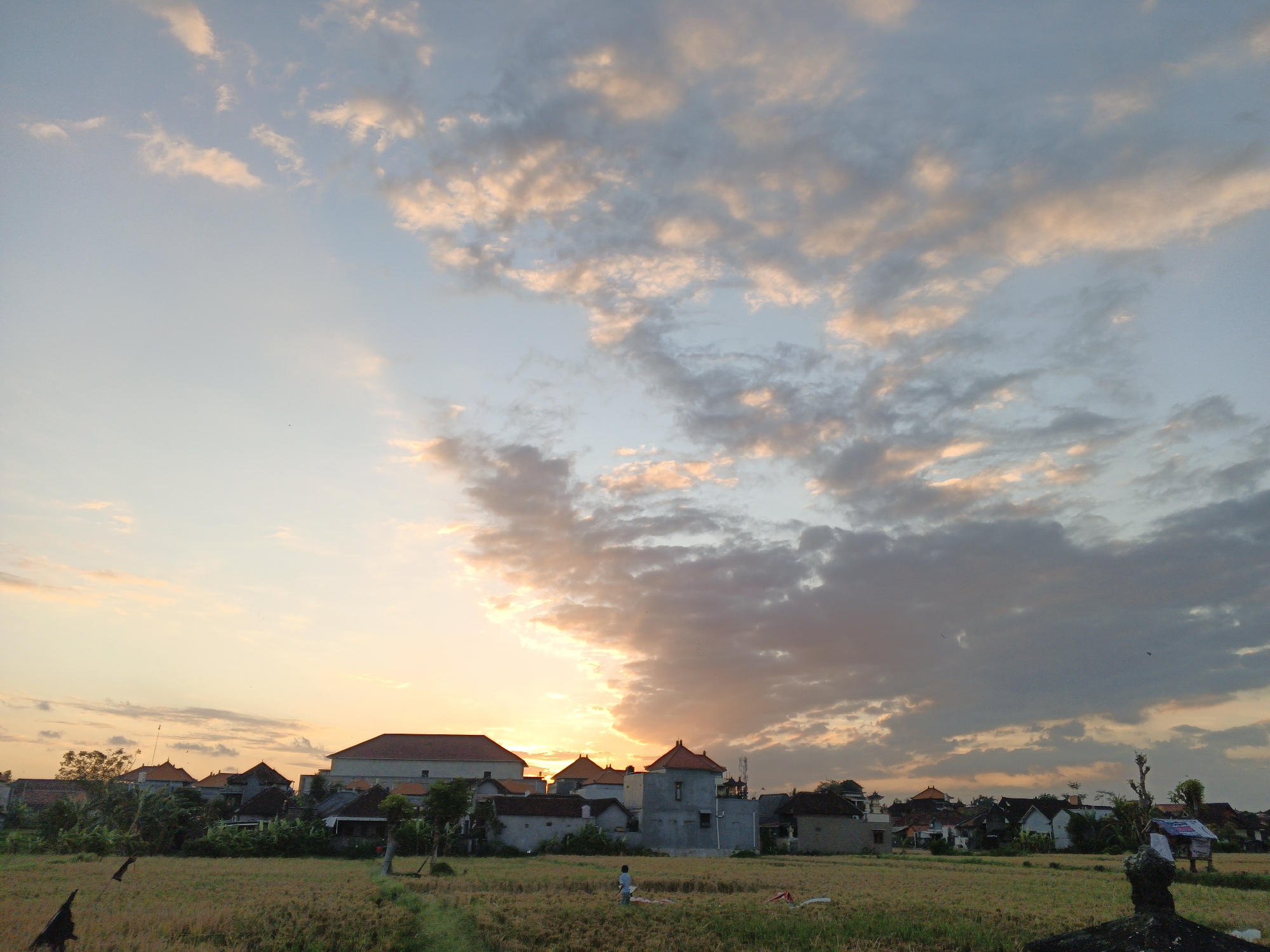 The image depicts a rural landscape at sunset in Sukawati, Indonesia. The sky is filled with a mix of pale blue and dramatic clouds tinged with orange and pink hues from the setting sun. In the foreground, a field of harvested or drying rice stalks stretches across the scene.
A small village is visible in the middle ground, characterized by buildings with terracotta-tiled roofs and various architectural styles. Some of the structures appear to be houses, while others might be community buildings or smaller shelters. The vegetation around the village is lush and green.
In the field, a solitary person, possibly a child or a young adult, stands in the center, facing away from the camera. The figure appears to be engaged in some activity, perhaps flying a kite or playing, though the specifics are unclear. There are also some dark, abstract shapes in the foreground that could be scarecrows or remnants of agricultural equipment.
The overall impression is one of tranquility and rural life at the close of day. The lighting suggests the golden hour, casting a warm glow over the scene. The image captures a slice of everyday life in a Southeast Asian countryside setting.