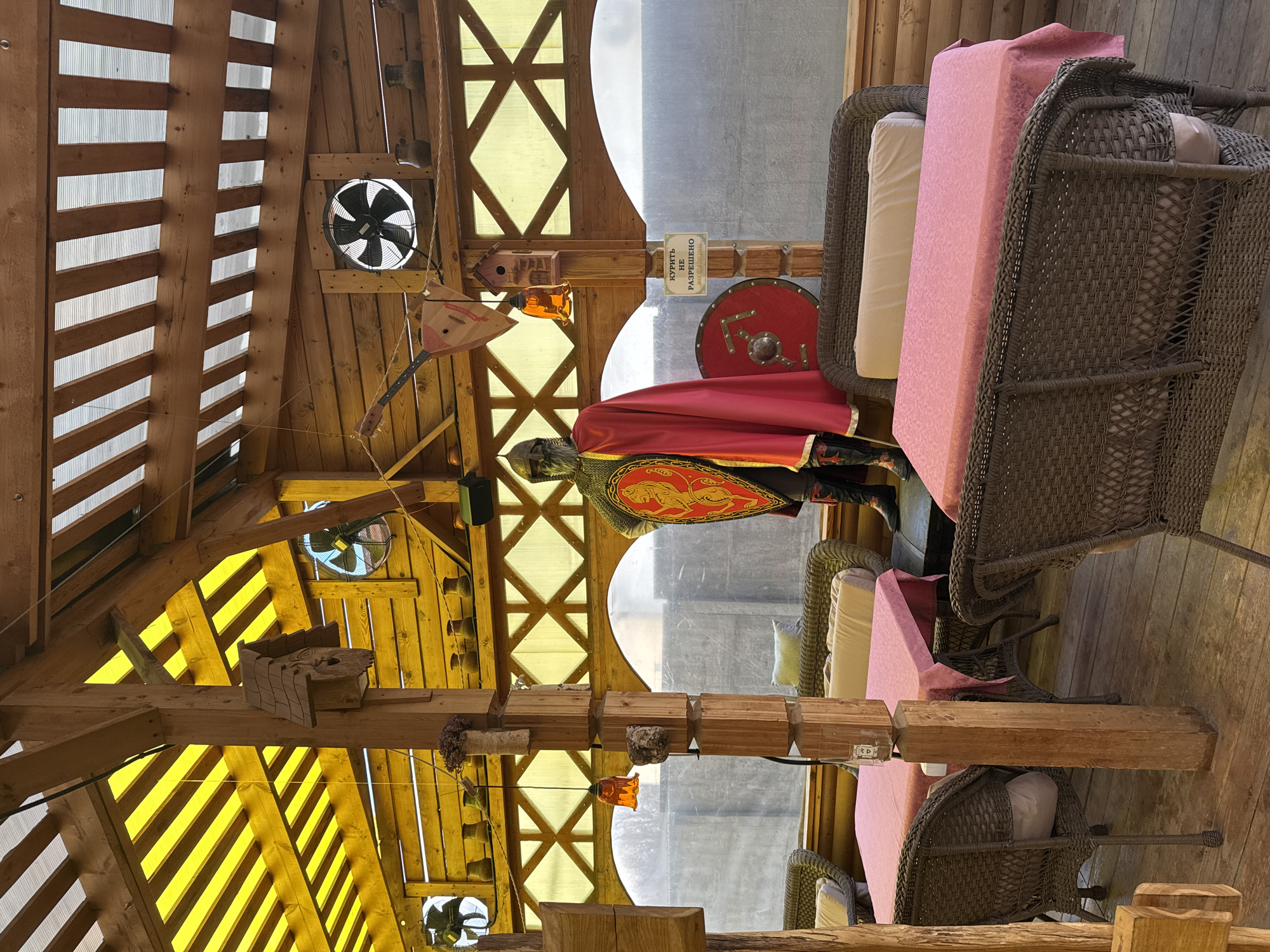 A restaurant in Kladovitsy, Russia, features a faux medieval theme. A suit of armor stands by the window, complete with a red cape, shield, and helmet. Wicker chairs surround tables with pink tablecloths. The wooden structure houses fans and decorative items such as birdhouses and musical instruments. A sign in Russian indicates that smoking is not allowed.