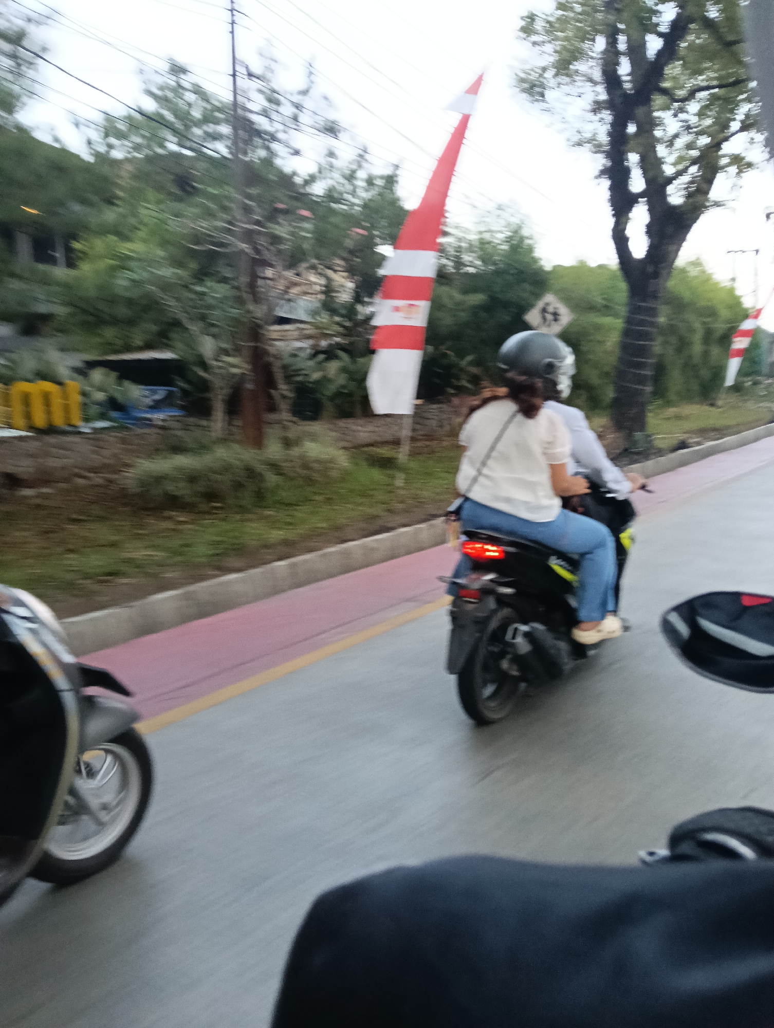 The image shows a scene on a street in Tasikmalaya, Indonesia, likely during the day. In the foreground, a motorcycle is partially visible on the left side of the frame, and the back of another rider or passenger is in the lower right. The central focus is a black motorcycle carrying two people. The rider, wearing a helmet, is operating the vehicle. The passenger, a woman wearing a white top and blue jeans, is seated behind the rider and looking to the left. The motorcycle has a visible taillight that is illuminated. The road appears to be paved, with a pink and yellow painted lane to the left of the motorcycle's path. In the background, there are trees, some foliage, and what appear to be buildings and utility poles. Two red and white flags, possibly Indonesian flags, are prominently displayed in the mid-ground, one tall and thin, and another shorter one partially visible. A traffic sign with a diamond shape and an image inside is also visible behind the motorcycle. The scene suggests movement, possibly on a road with moderate traffic.