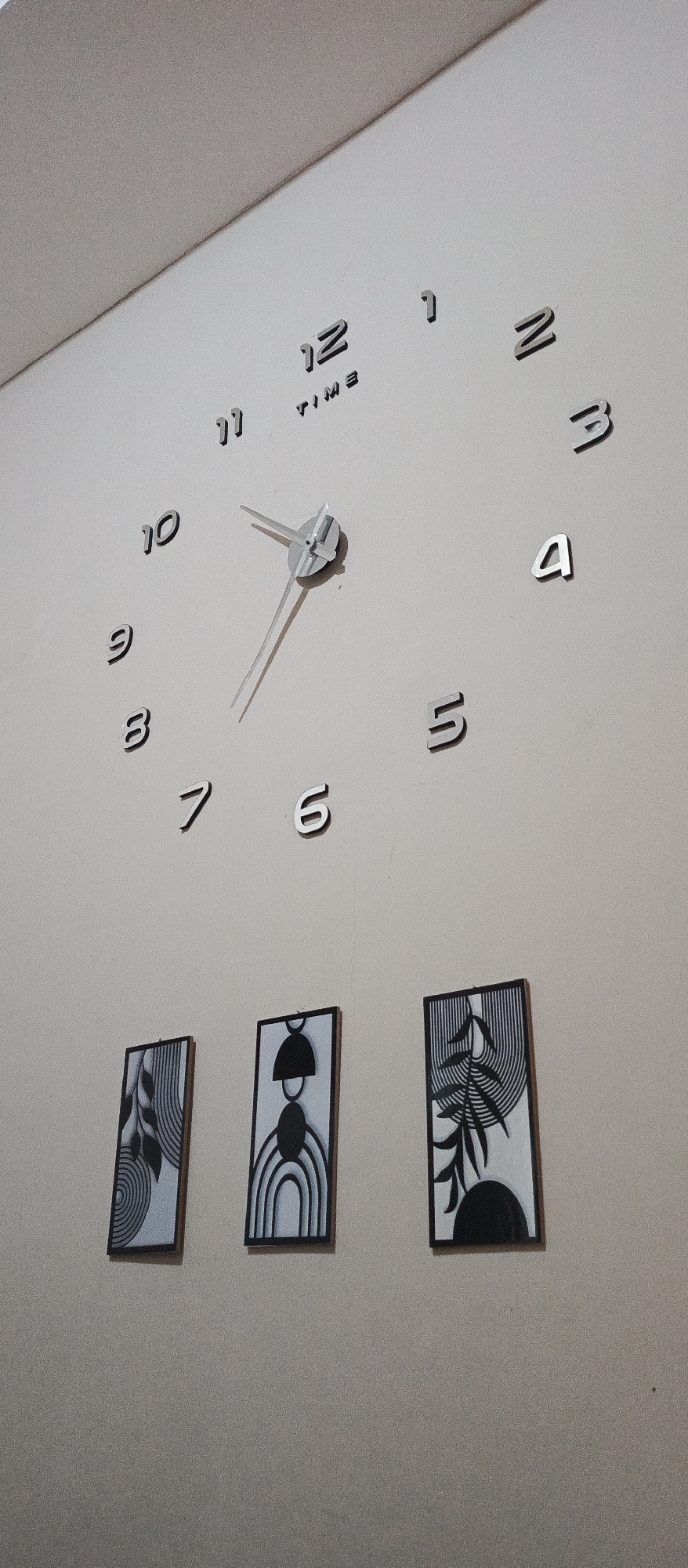 A modern, minimalist wall clock with silver numbers and hands is mounted on a light-colored wall. The clock face is not visible; instead, the individual numbers (1-12) are placed directly on the wall in a circular arrangement. The hour hand points to approximately 10, and the minute hand points to approximately 4. The word "TIME" is printed in small black letters below the number 12. Below the clock, three vertical rectangular pieces of abstract art are hung. Each artwork features black and white geometric patterns. The art on the left shows concentric circles and leaf-like shapes. The middle artwork has a prominent semi-circular arch shape with radiating lines. The artwork on the right depicts a stylized plant with linear patterns. The background appears to be a plain wall with a slight texture. The image was taken in Jatisari, Indonesia.
