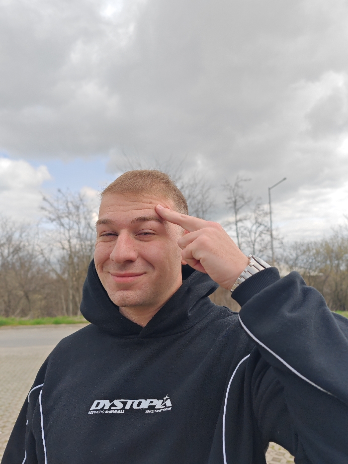 One adult male is depicted in an outdoor setting under a partly cloudy sky. The male subject has light skin, a buzz cut with light brown hair, and appears to be smiling while looking directly at the camera. He is wearing a black hooded sweatshirt with white piping along the sleeves and sides. The sweatshirt features white text on the chest that reads "DYSTOPIA AESTHETIC AWARENESS SINCE NINETYNINE." His left arm is raised, with the index finger of his left hand pressed against his forehead above his right eye. A silver-colored watch with a metallic link bracelet is visible on his left wrist. The background consists of bare deciduous trees, suggesting late winter or early spring conditions, and a paved surface in the lower left. A street light or utility pole is visible in the upper right background. The specific location within the city of Shumen, Bulgaria, cannot be determined from the visual information provided in the image.