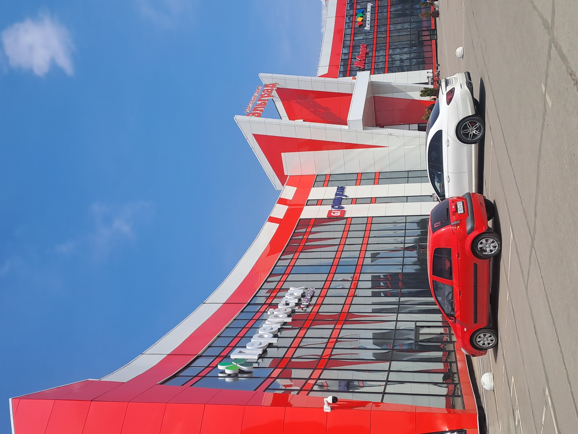 The image shows the Elektrostal shopping center, located in Elektrostal, Russia. The building is primarily red and white with large reflective windows. "Эльград" is written in Russian on the upper section of the building. A red Hyundai Getz and a white sedan are parked in the parking lot in front of the shopping center. The sky is bright blue with a few clouds.