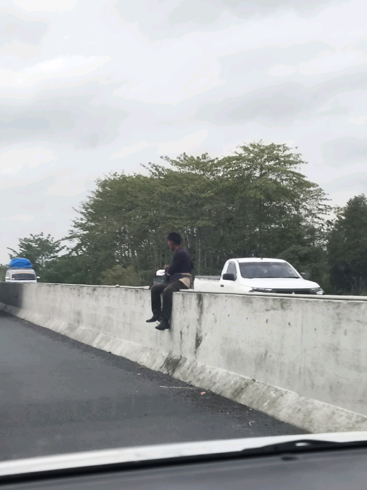 A person is sitting on a concrete barrier next to a highway. The person is wearing dark clothing and black shoes. They are looking to their left, away from the camera. There is a white pickup truck driving on the highway behind the person, and a white van with a blue tarp on top is visible further down the highway. Lush green trees line the background. The sky is overcast. The scene appears to be in a rural or semi-rural area, given the presence of trees and the absence of dense urban development. The highway barrier suggests the location is a major roadway, possibly an expressway or a highway bypass. The area is identified as being in Pematang Kijang, Indonesia.
