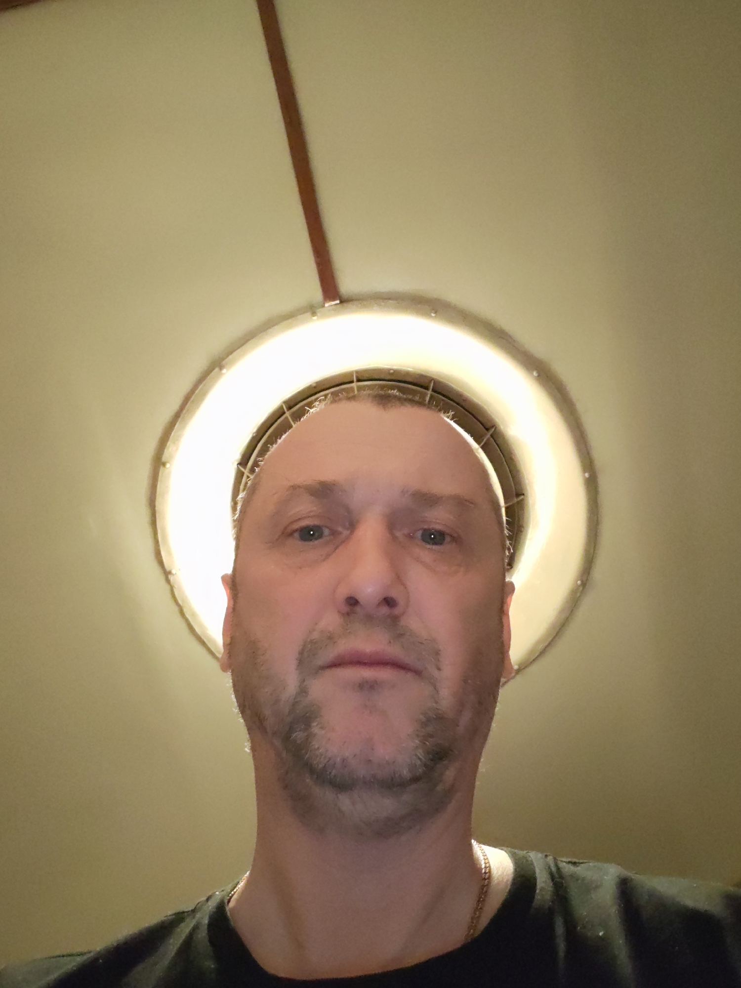 This low-angle selfie, taken indoors in Shcheleiki, Russia, features a middle-aged man. His face and upper torso are visible, filling the lower portion of the frame. He has short, light-colored hair with a receding hairline and a light beard or stubble. His eyes are blue or grey, and he maintains a neutral expression. He is wearing a dark, presumably black, t-shirt, with a thin gold-colored chain visible around his neck.
Directly behind the man's head, mounted on a light-colored ceiling, is a brightly illuminated circular light fixture, creating a halo effect. A thin, dark, linear element, possibly a ceiling beam or wire, diagonally traverses the upper part of the ceiling, touching the top edge of the light fixture. The ambient lighting in the room appears warm. No discernible text is visible in the image. The perspective suggests the photo was taken with the camera held low, pointing upwards towards the subject and the ceiling.