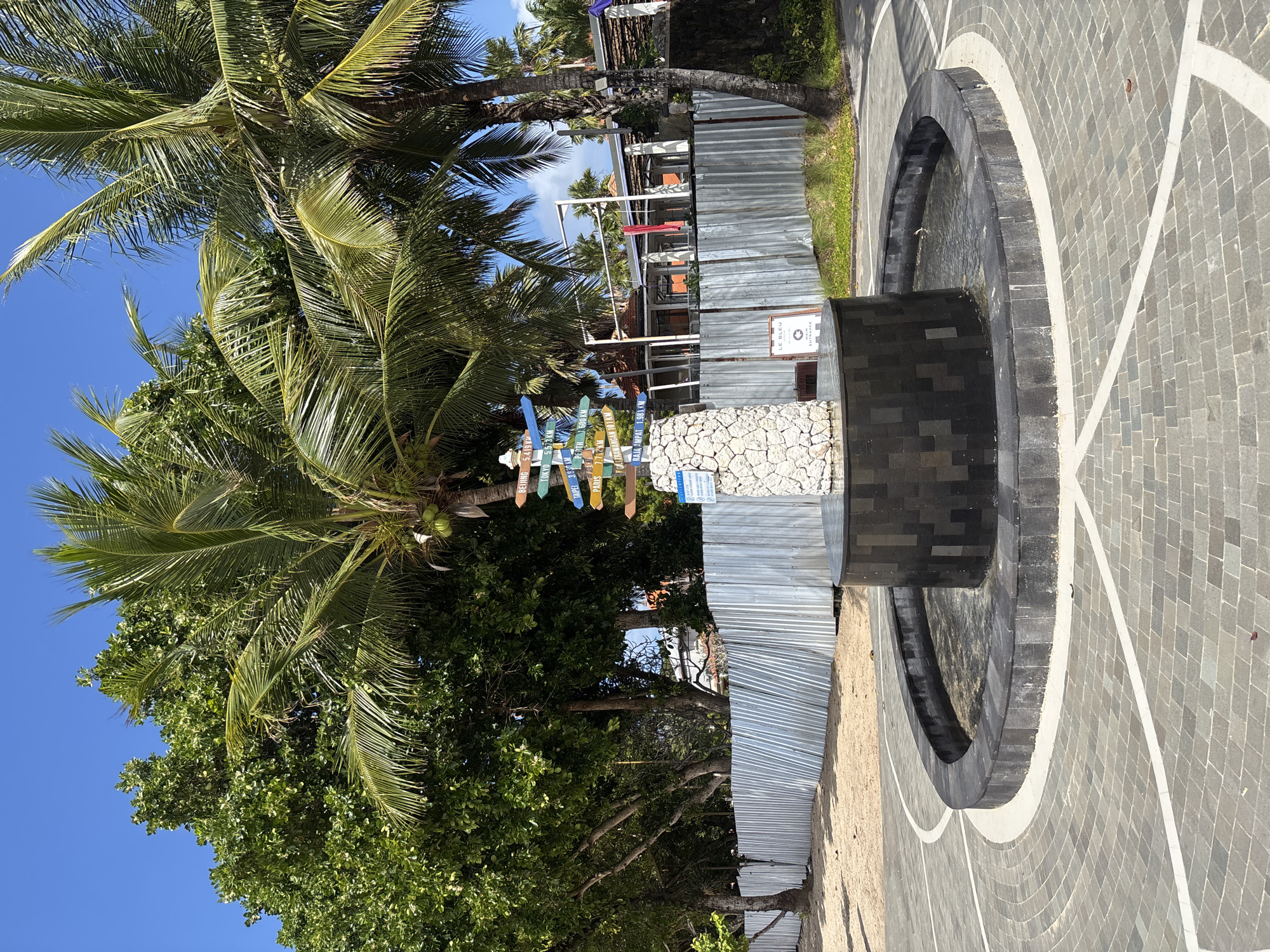 This is a vertical, eye-level shot of a tropical outdoor scene. The foreground features a circular, dark stone fountain with a section of water visible, surrounded by a paved area with a grey cobblestone pattern and white curved lines. To the left of the fountain, there is a signpost with multiple colorful directional signs pointing to various locations and distances, such as "BEIJING 5416 KM," "TOKYO 5373 KM," "NY 3931 KM," "PARIS 5851 KM," and "RAJA AMPAT 1981 KM." Behind the signpost, there are palm trees and lush green foliage under a clear blue sky. A corrugated metal fence runs horizontally across the mid-ground, partially obscuring a building with a light-colored exterior and some red accents visible on its upper level. A small sign on a stone base near the fence reads "LE BLEU BEACH CLUB ENTRANCE." The overall scene suggests a sunny day in a tropical location, likely a beachside resort or public area. There are no people visible in the frame. The presence of directional signs to far-off cities indicates a popular tourist destination.