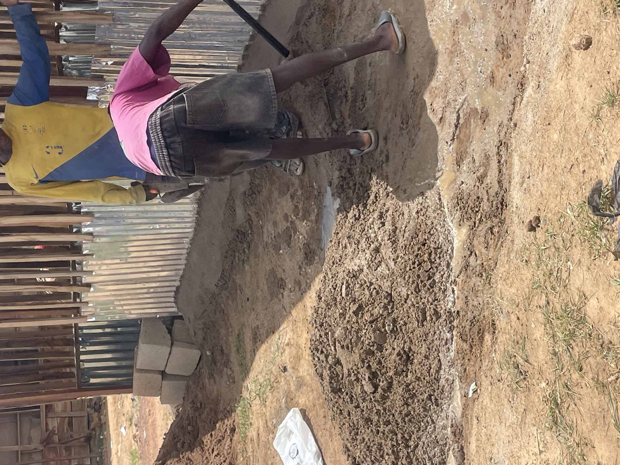 In Durimi, Nigeria, two individuals are working on a construction site. One person is wearing a yellow and blue long-sleeved shirt, while the other has on a pink shirt and grey shorts. The second individual is holding a black pole and appears to be mixing a pile of dirt with water to form mud. The ground is a mix of dirt, mud, and grass. There is a building under construction with corrugated metal sheets for walls. Cement blocks are seen stacked near the base of the building. A white plastic bag is on the ground nearby.