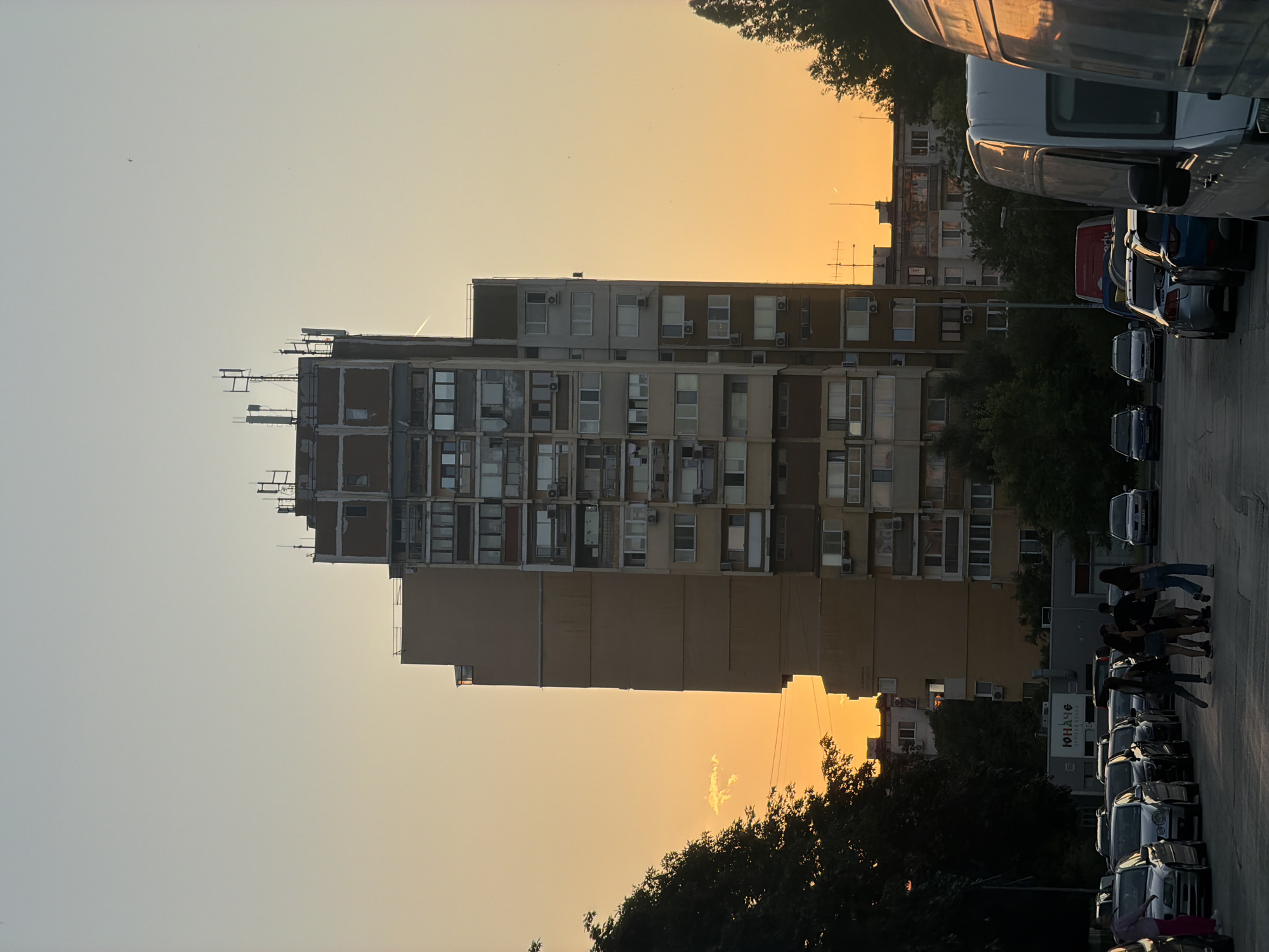 The image captures an urban scene in Sofia, Bulgaria, during what appears to be sunset, given the warm hues of the sky. The focal point is a tall, Brutalist-style apartment building with a weathered facade, featuring numerous windows and balconies. The building's exterior is a mix of beige and grey concrete with brown accents around the window frames. On its roof, several telecommunication antennas are visible.
In the foreground, a line of parked cars stretches down a paved road, with several vans partially obscuring the view on the right side. The vehicles are varied in make and model, suggesting a typical urban parking area.
A group of six young people, three women and three men, are walking together on the road, silhouetted against the brighter sky. They are casually dressed and appear to be engaged in conversation.
To the left of the frame, a dense canopy of green trees partially obscures another building and the setting sun. On the right, more trees are visible, their dark foliage contrasting with the illuminated sky.
A sign with Cyrillic lettering, "ЮНАЧЕ" (YUNACHE), is mounted on a low-rise building in the middle ground, possibly indicating a business or establishment. The overall atmosphere is one of a typical evening in a residential district of Sofia, characterized by apartment living, parked vehicles, and the presence of people in a public space.
