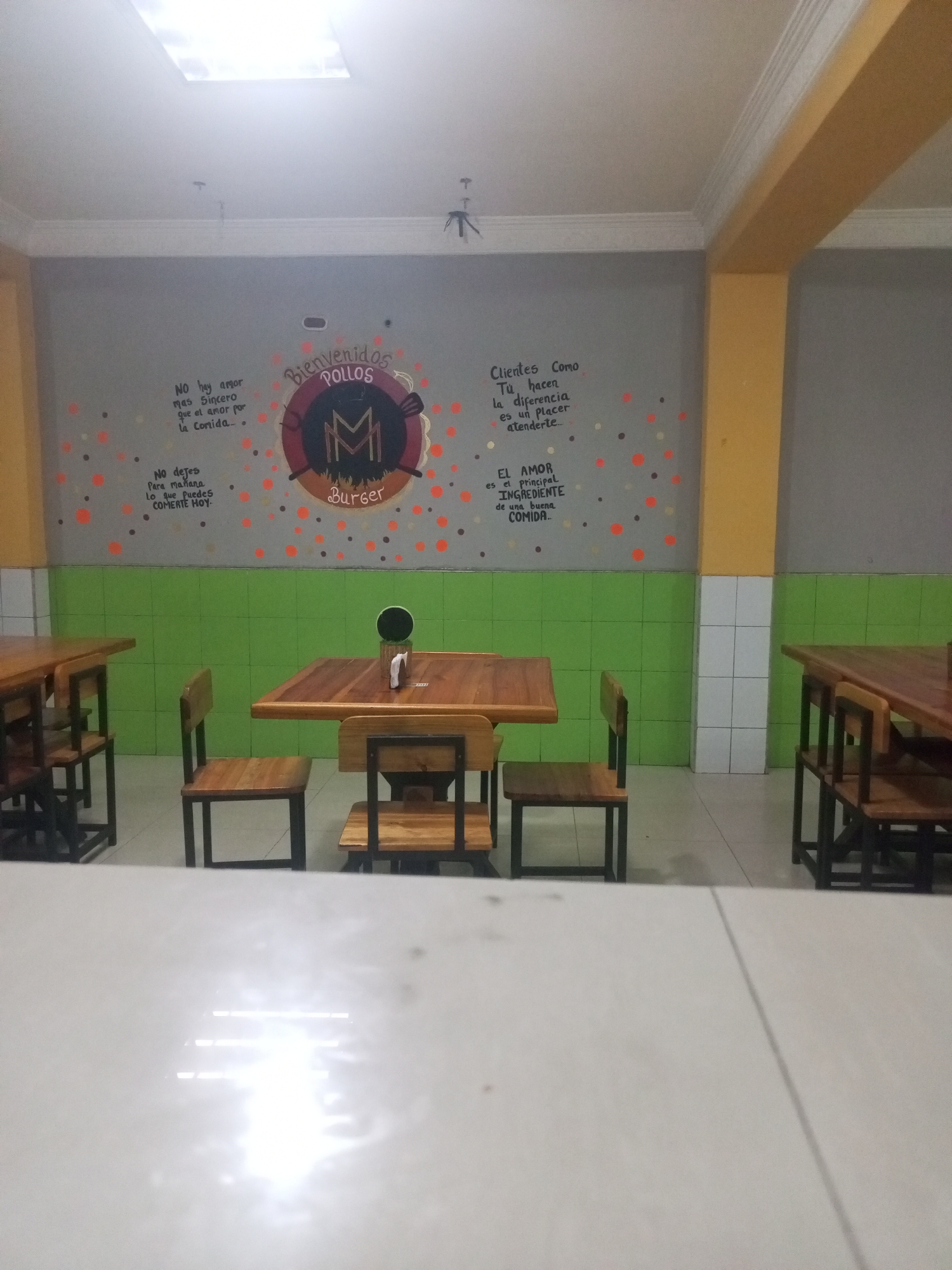 This appears to be an interior shot of a restaurant in Juan Griego, Venezuela. The decor includes a mural with the words "Bienvenidos Pollos" and a logo of the letter M within a circle, followed by the word "Burger." The mural also includes scattered dots of orange, red, and brown. Adjacent to the mural are phrases about food, such as "No hay amor mas Sincero que el amor por la Corrida..." and "EL AMOR es el principal INGREDIENTE de una buena COMIDA..."
The restaurant has tables and chairs, primarily made of wood. The walls are painted gray with a green tiled area. The lower part of some walls is covered in white tiles. The overall lighting is bright.