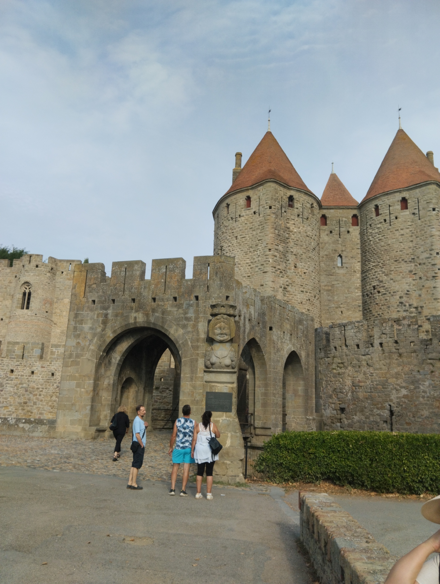 The image depicts the entrance to the Cité de Carcassonne, a fortified medieval city located in Carcassonne, France. The foreground shows a paved walkway leading towards a large stone archway, part of the city's defensive walls. Several individuals are visible. Two men and a woman stand near the archway, appearing to observe the structure. Another woman walks away from the camera towards the arch. Behind them, the stone walls of the Cité rise, characterized by their aged, weathered appearance. The walls are punctuated by arched openings and battlements. The sky above is overcast with a pale, cloudy sky. The architecture suggests a historical and defensive structure.
