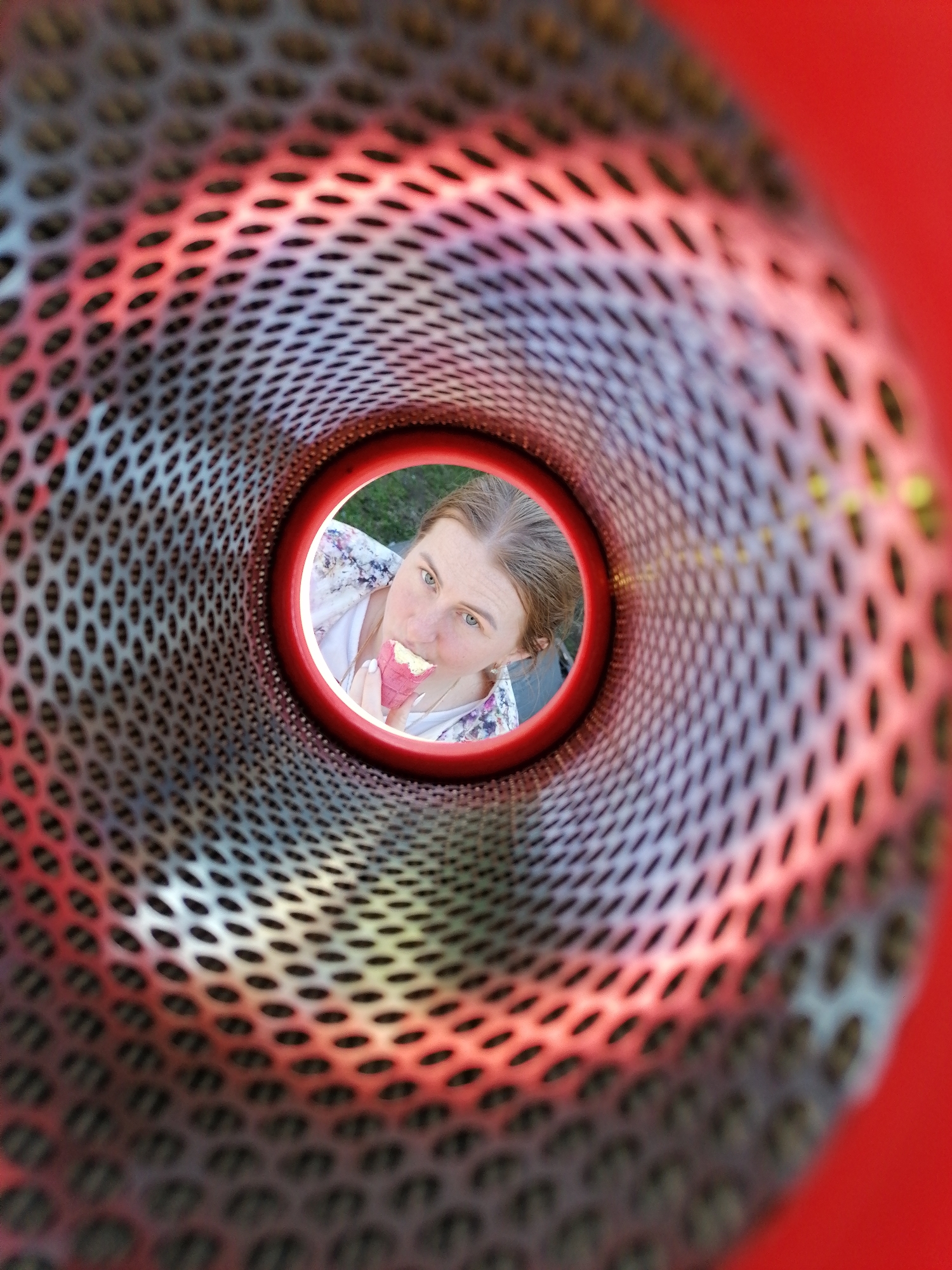 A woman is visible through a circular opening in what appears to be a metal structure with numerous small holes, possibly part of playground equipment. The structure is primarily red and silver, and the woman is eating an ice cream cone with a pink cone. The background behind the woman suggests an outdoor setting with greenery. The location is Unknown, Unknown.