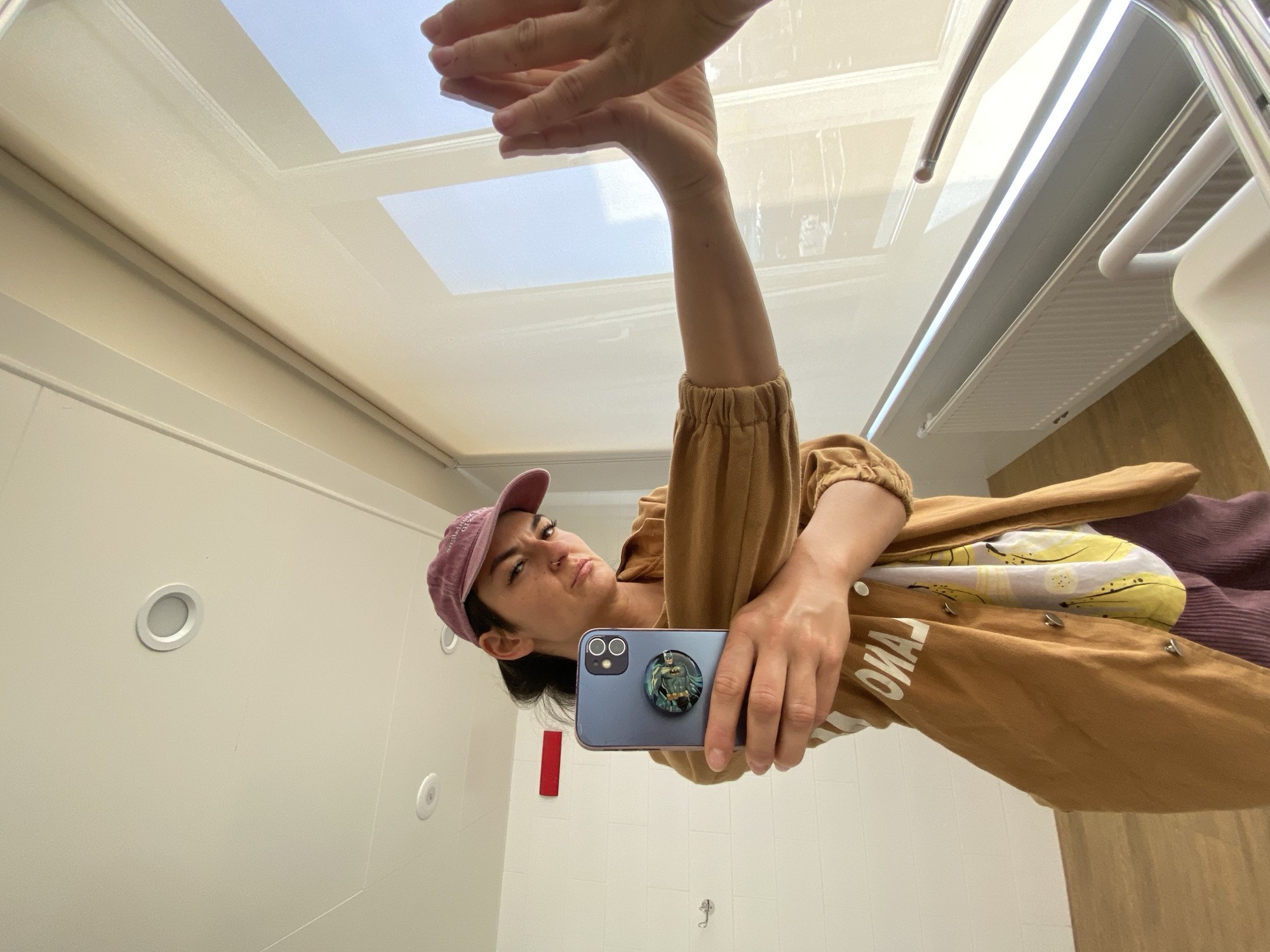 A reflection in a mirror shows a person holding a phone, wearing a pink baseball cap and a tan jacket. The environment appears to be an indoor space, possibly a bathroom or restroom, with white walls and ceiling. Features visible in the reflection include recessed lighting in the ceiling and a skylight. The phone has a pop socket with a Batman design. The location is not discernible from the image.