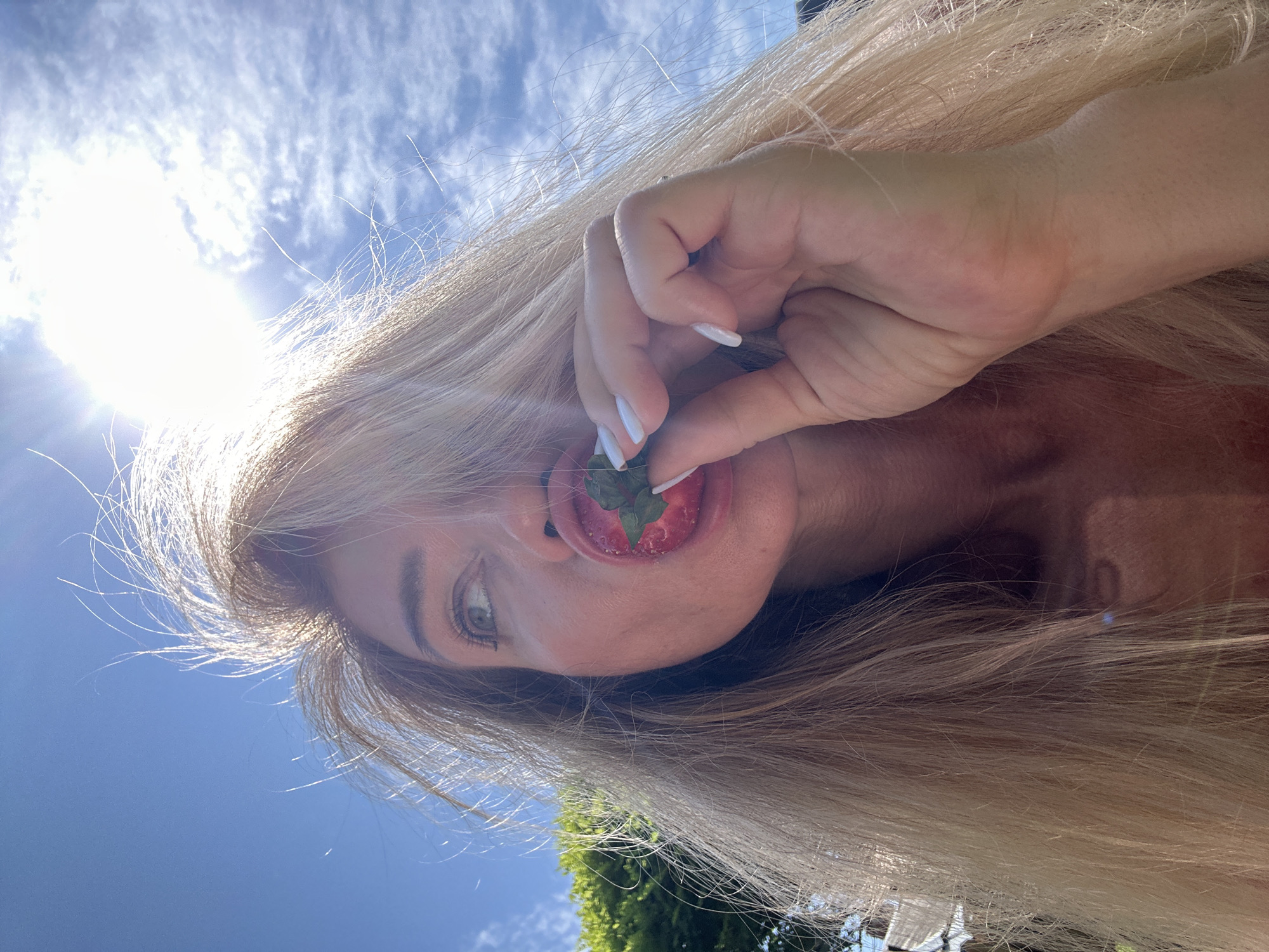 A close-up, low-angle selfie shows a woman's face and her hand holding a strawberry to her mouth against a bright blue sky with scattered white clouds. The sun is shining intensely, creating a lens flare in the upper right portion of the image and backlighting the woman's long, blonde hair, making strands appear to float. She has light-colored eyes and is looking slightly upwards and to her left. Her lips are red, and she is holding a strawberry with its green leaves, about to take a bite. Her fingernails are painted white. The background shows a blurred green tree line on the left and some indistinct structures. The image has a candid, outdoor feel, suggesting a warm, sunny day. The location in Armavir, Russia, is not visually discernible from the image itself.