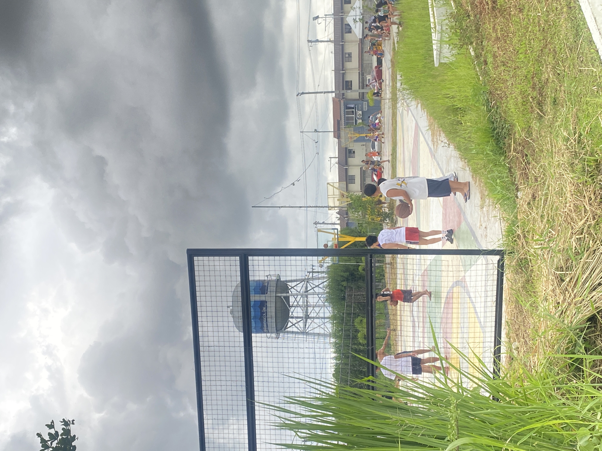 The image depicts an outdoor basketball court in what appears to be a residential area. Several individuals are present on and around the court, some actively playing basketball while others observe. The court itself has a colorful, geometric pattern. In the background, there are houses, utility poles, and a prominent water tower that reads "Davao Water Corporation's, Inc." The sky is overcast with heavy, dark clouds, suggesting an impending storm or rain. Lush green grass and vegetation are visible in the foreground and around the court. The presence of the water tower specifically identifies the location as Davao City, Philippines.