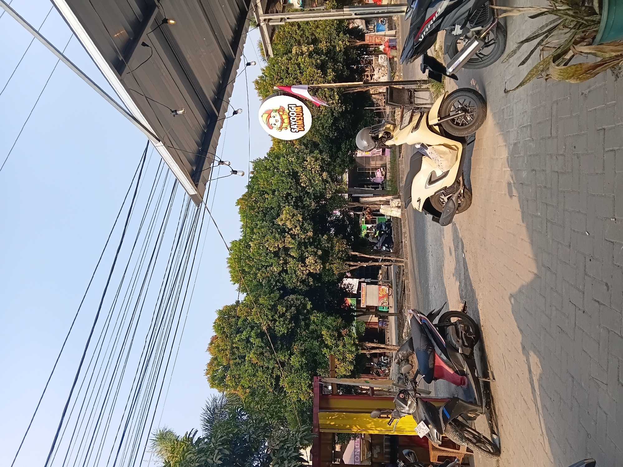 In the city of Wibawamulya, Indonesia, a roadside scene unfolds under a clear, bright sky. Multiple black electrical wires crisscross the upper portion of the frame, casting thin shadows. A corrugated metal awning extends from the right side of the image, supported by a silver metal beam, and strung with several bare lightbulbs.
Below the awning, a round, white sign with a cartoon character and the text "SAYOOGI DIMSUM" is illuminated. A small Indonesian flag flutters from a pole next to the sign. The foreground is paved with interlocking brick pavers, upon which three motorbikes are parked. The leftmost motorbike is black, with a red seat cover and a license plate visible. The middle motorbike is cream-colored, with a black seat and a helmet resting on it. A shopping bag is attached to the front of this motorbike. The rightmost motorbike is black, with a silver exhaust pipe.
Behind the motorbikes, a street is visible with more trees lining the other side. A yellow and red structure stands on the left, possibly a food stall. Further down the street, other buildings and more trees can be seen. The overall atmosphere is that of a casual, possibly commercial area during daylight hours, with shadows indicating the sun is at a moderate angle. No people are clearly visible in the image, though there are hints of activity in the background. A potted plant with dried leaves sits in the lower right corner of the frame.