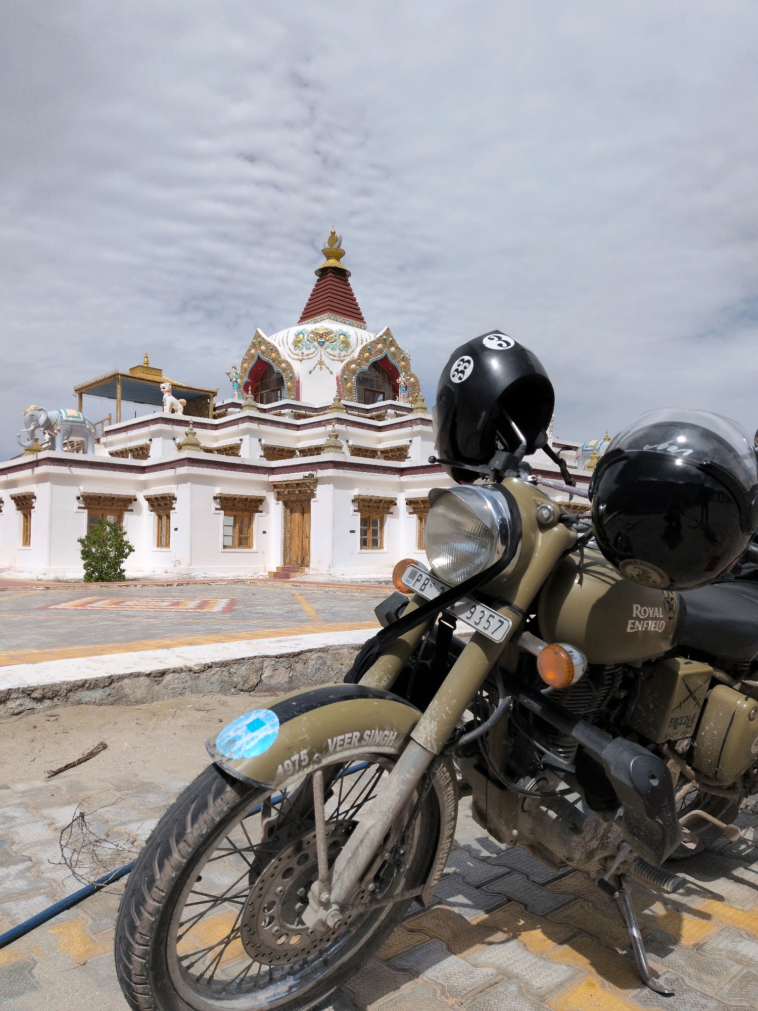 An outdoor, eye-level, medium shot captures a Royal Enfield motorcycle parked on a paved surface in front of a white Buddhist stupa in Martselang, India. Two black helmets are attached to the handlebars, one perched on top of the other. The motorcycle, a sandy khaki color, is positioned diagonally with its front wheel closest to the viewer. The stupa, a whitewashed structure with red and gold ornamentation, stands prominently in the background. The sky above is overcast with scattered clouds. The foreground is a mix of sandy and paved ground with some debris. The license plate of the motorcycle is partially visible, displaying "PB 7 9357". The word "VEER SINGH" is visible on the fender of the motorcycle.