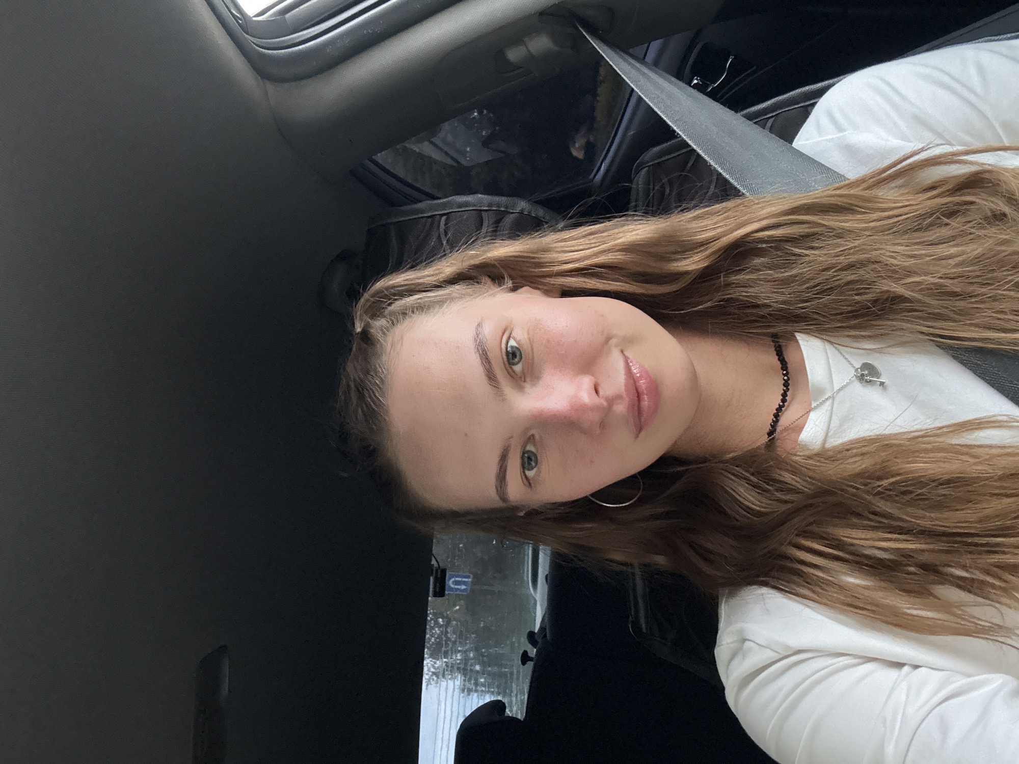 A young woman with long, wavy blonde hair tied in two pigtails is in the driver's seat of a car. She is wearing a white long-sleeved shirt and a black choker necklace with a silver pendant shaped like a key. Her left ear is adorned with a silver hoop earring. The woman has light blue eyes and fair skin with a slight flush on her cheeks. She is looking directly at the camera with a subtle smile. A gray seatbelt is visible across her chest. The interior of the car is dark, with the passenger seat and the ceiling of the car visible. Through the car windows, trees and a street sign with a "U" shape can be seen, suggesting an outdoor location. The lighting appears to be natural, possibly from an overcast day. The image does not provide enough visual information to determine the specific location within the city of Perm, Russia.
