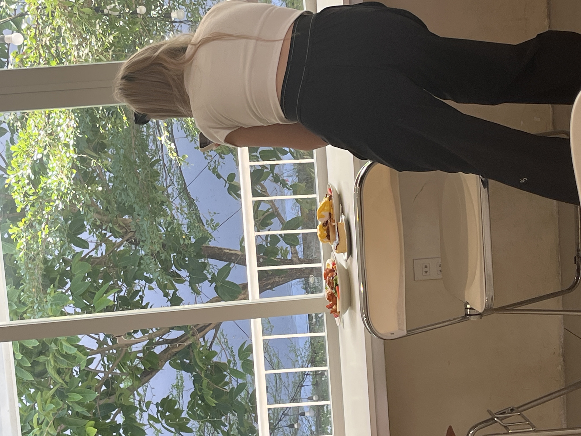 A blonde woman in a white t-shirt and black pants is looking at food on a table near a window. The window is framed by a white grid and reveals green trees and a blue sky. A clear chair with silver legs sits next to the table. A dual-outlet power socket is visible on the wall near the chair. The location is likely in Đà Nẵng, Vietnam, based on the vegetation and architecture.