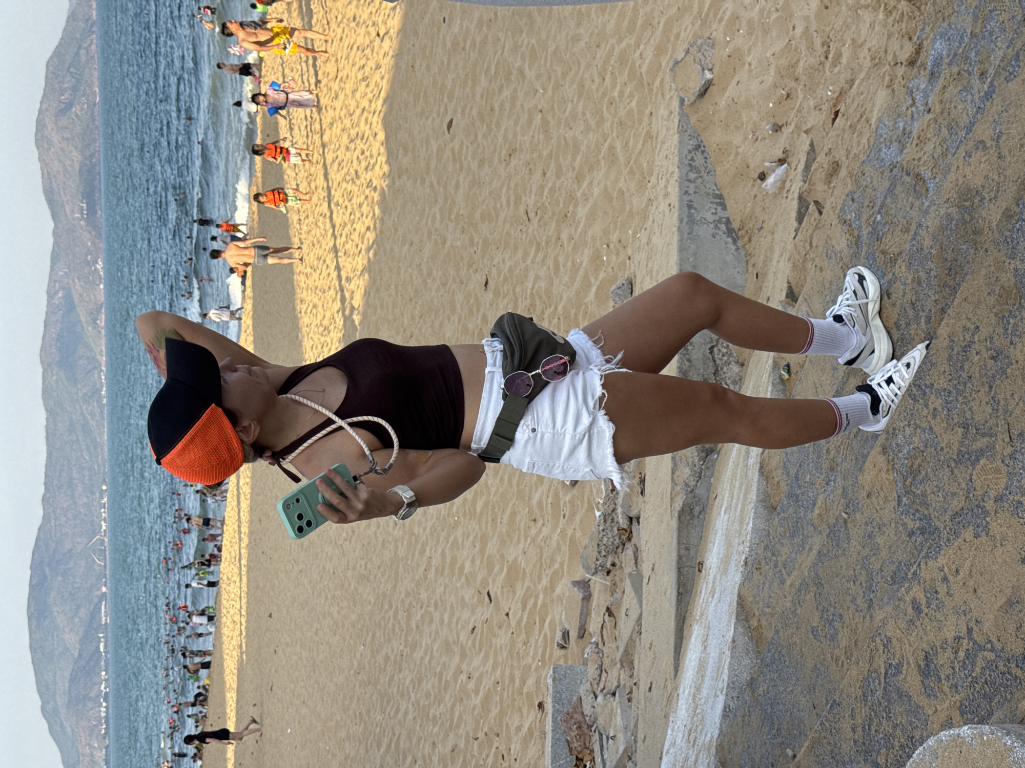 A young woman stands on a sandy beach near the city of South Quy Nhon Ward, Vietnam, holding a smartphone. She is wearing a dark crop top, white denim shorts, a green fanny pack, white socks with red stripes, and athletic shoes. She also wears a black and orange baseball cap and sunglasses are clipped to her fanny pack. The beach is crowded with people swimming and walking in the ocean and on the sand. In the background, mountains are visible. The woman appears to be looking out at the ocean.
