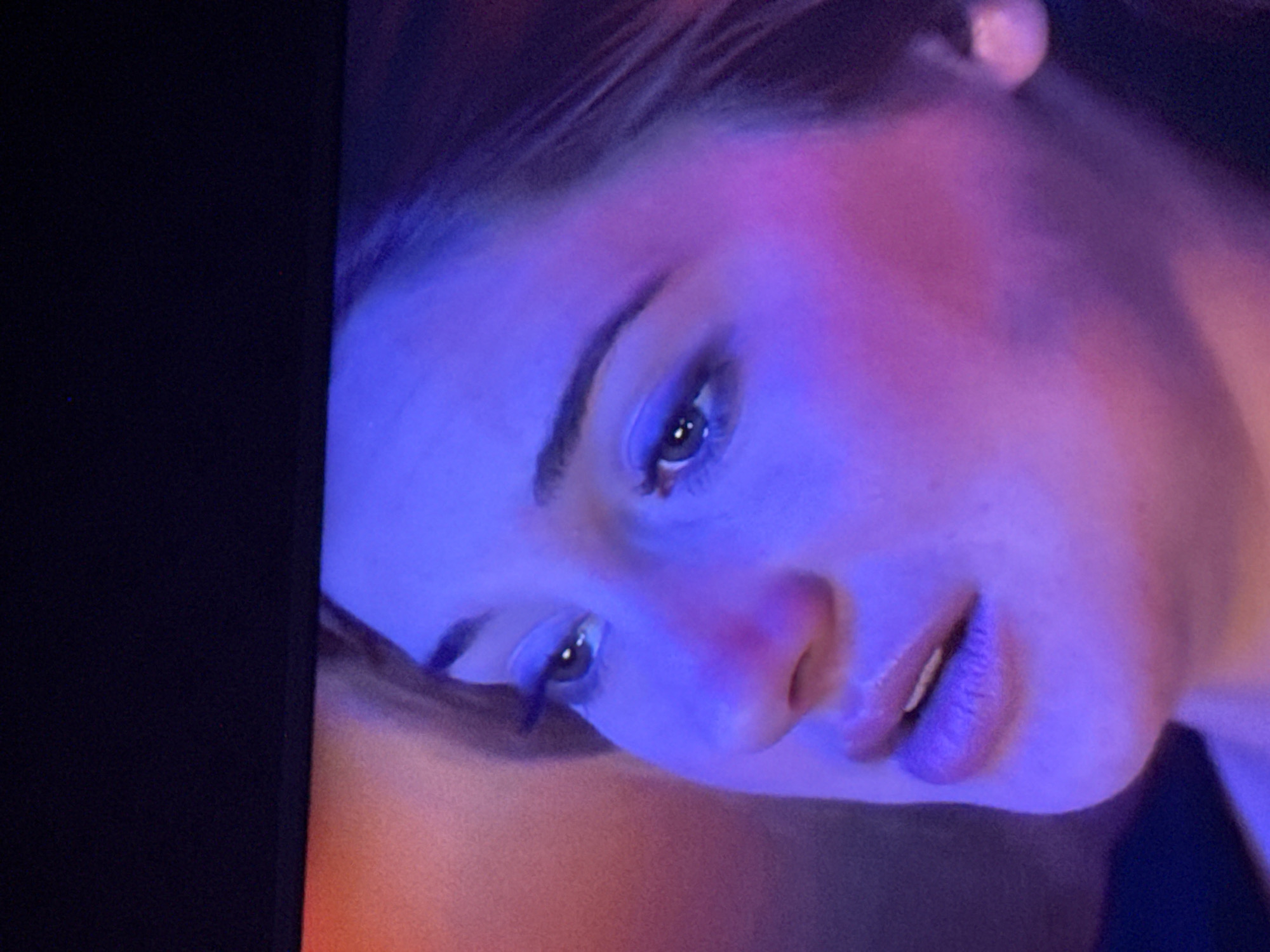 A close-up, low-angle shot of a woman's face. Her face is illuminated with a blue and pink light. Her eyes are partially open and focused downward. The left side of the image is black, possibly indicating a dark background or edge of a screen. The location and city are not discernible from the given image.