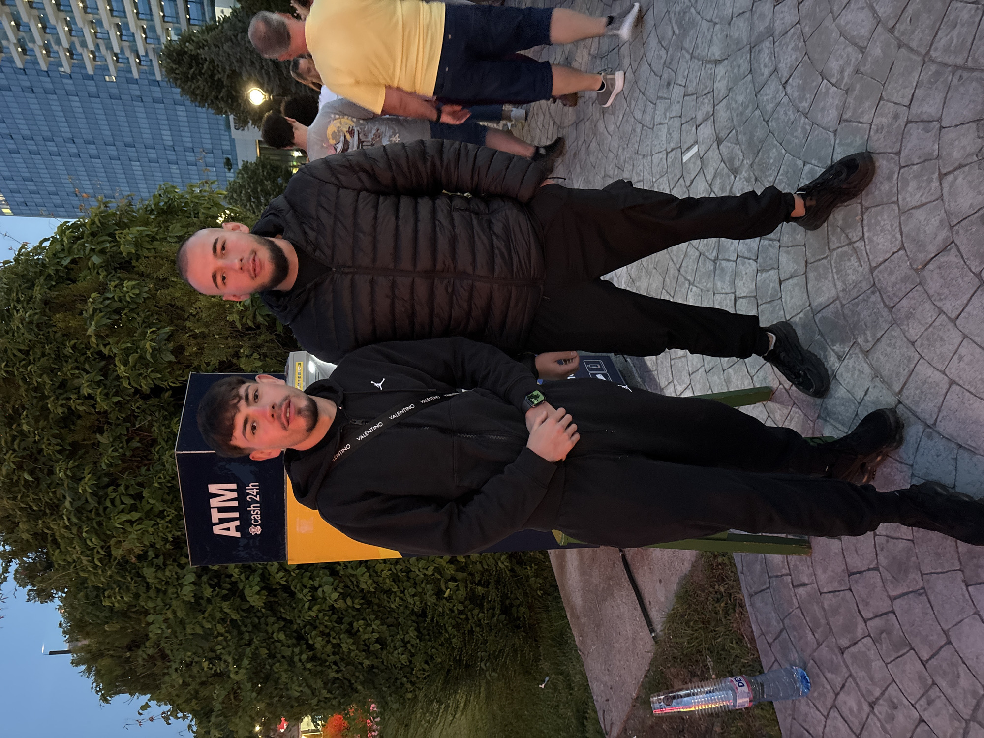 The image depicts an outdoor, urban scene at dusk or early evening, identified as the city of Varna, Bulgaria, based on the presence of a "DEVIN" brand water bottle.
In the foreground, two young adult males stand facing the camera. The male on the left has dark hair with a fringe, slight stubble, and wears a black Jordan-branded full-zip hoodie, black Jordan-branded jogging pants, and black athletic shoes with translucent soles. He has a black Valentino-branded shoulder bag strap across his chest and a green digital watch on his left wrist, with his hands clasped in front. The male on the right has a shaved head, a short dark beard, and wears a black puffer jacket, black cargo pants, and black athletic shoes. He stands with his hands at his sides or in pockets.
Between the two subjects, slightly to the left, is a dark blue and yellow ATM machine displaying "ATM" in white capital letters and "cash 24h" with a currency symbol below it. To the lower left, on the grey/tan patterned paving stones, a clear plastic bottle of "DEVIN" mineral water lies on its side.
In the background, lush green bushes and trees create a dense screen behind the ATM and extending to the left. To the right, modern multi-story buildings with numerous windows and balconies are visible against a twilight blue sky. Several other individuals are present in the mid-ground and background, including a man in a yellow t-shirt and dark blue shorts facing away, and another person in a grey t-shirt with a graphic design. A streetlamp emits a yellow glow in the mid-ground to the right. The ground is paved with interlocking stones arranged in a semicircular pattern.
