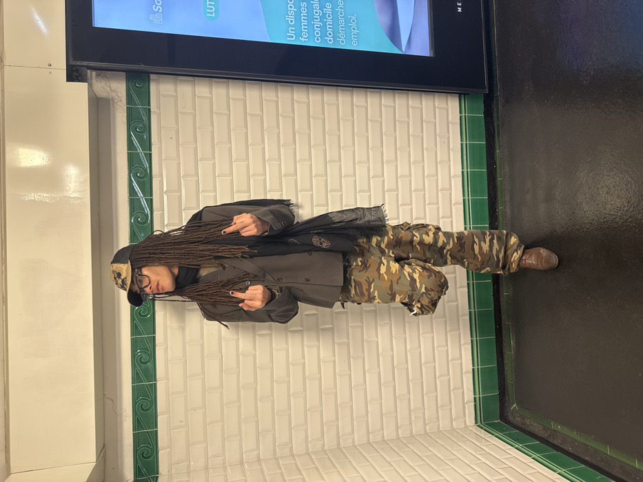 In a subway setting, a person with dreadlocks is photographed giving the middle finger with both hands. They wear a baseball cap, glasses, a dark jacket, and camouflage pants, with brown shoes completing the outfit. The backdrop consists of white tiled walls, dark flooring, and a digital advertisement screen above.