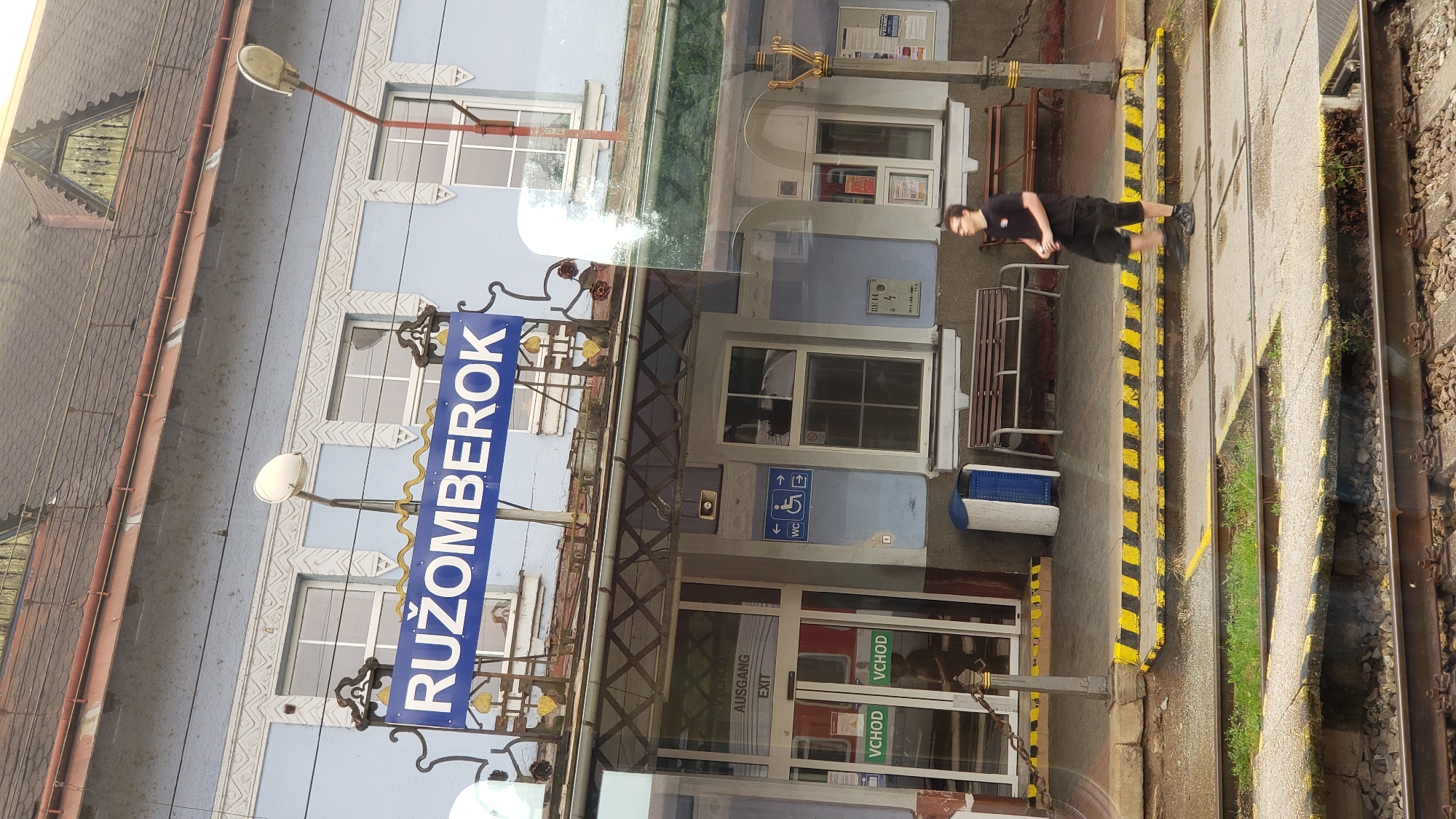 The image shows the exterior of the Ružomberok train station in Slovakia. A blue sign above the entrance reads "RUŽOMBEROK". The building is light blue with decorative white trim. There are several windows and doors. To the left of the entrance, there are signs indicating "VCHOD", "AUSGANG", and "EXIT". To the right of the entrance, there are signs for "WC" and a wheelchair accessibility symbol. A young man in a black t-shirt and shorts stands on the platform, holding a cup. Benches are visible on the platform. The platform has yellow and black striped safety markings. Train tracks are in the foreground.