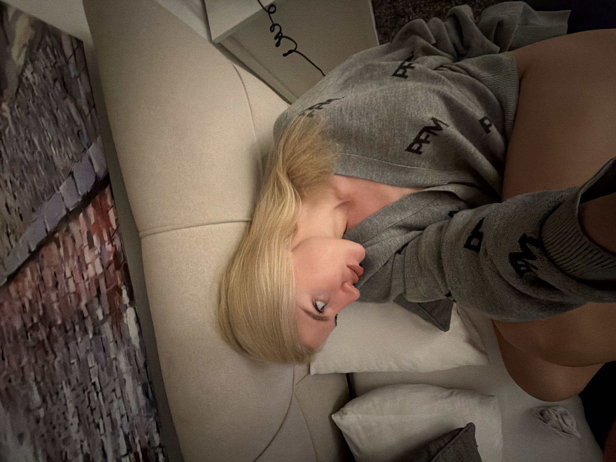 A high-angle, close-up photograph taken in an interior room in Belgrade, Serbia, depicts a blonde female reclining on a bed. The subject has long, straight blonde hair and fair skin, with her gaze directed towards the right side of the frame and her lips slightly parted. She is wearing a grey V-neck cardigan patterned with repeated black capital letters "PFM." Her upper thighs are visible below the cardigan. She is positioned against a light-colored upholstered headboard or bed frame, with two white pillows visible to her left. The wall above the headboard features a large wall art or wallpaper depicting a brick-like pattern in shades of grey, brown, and muted red. A black coiled cable rests on a partially visible white surface to the right of the bed. The scene is illuminated with dim, ambient light.
