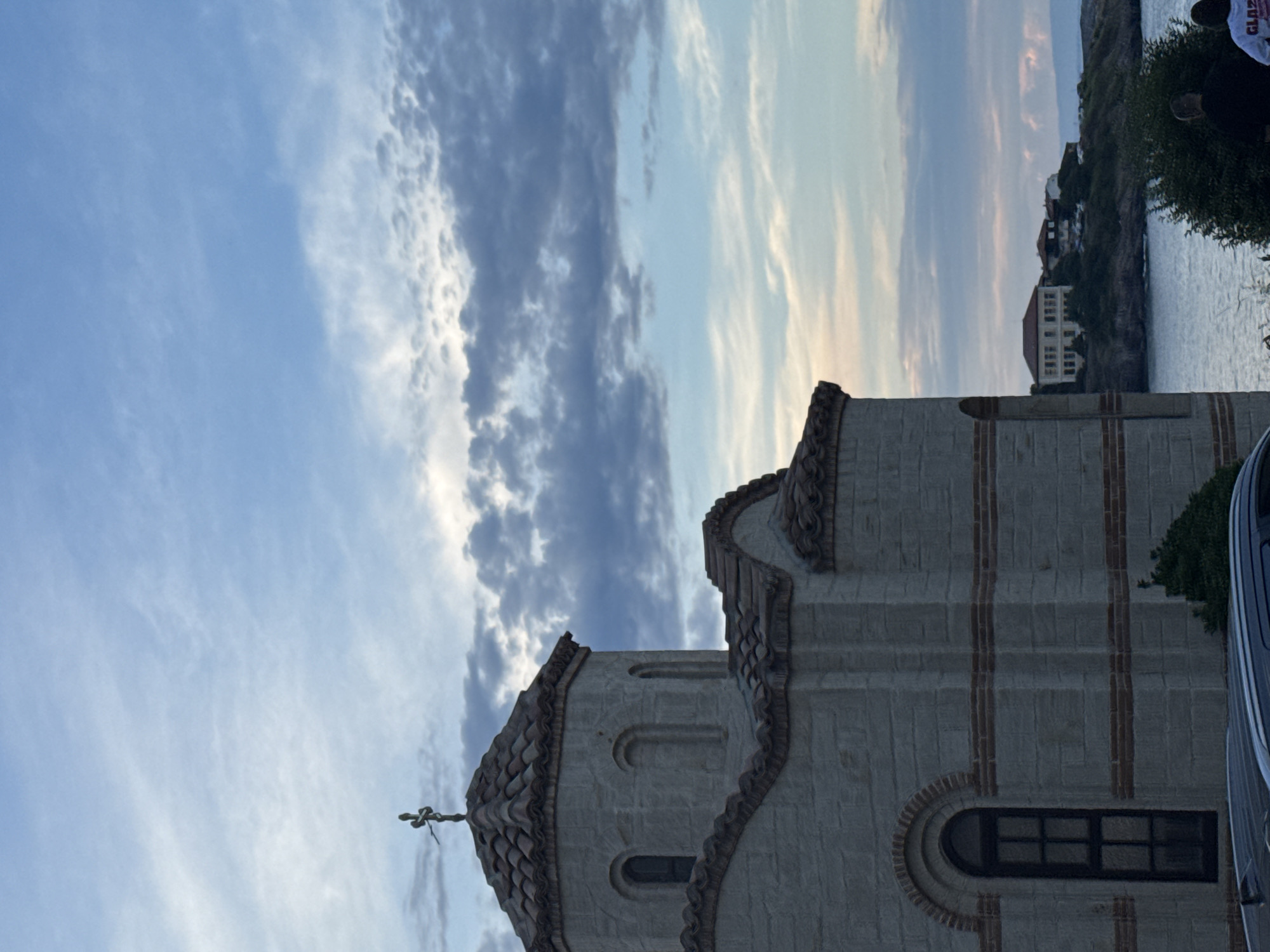 The image shows a stone church with a tiled roof and a cross on top, located in Sozopol, Bulgaria. The church is situated on a hill overlooking the sea, with several houses visible in the background on a nearby peninsula. The sky is filled with dramatic clouds, suggesting either sunrise or sunset. In the foreground, the roof of a car is visible, and two people are standing near the water, facing away from the camera. The overall scene depicts a coastal town in Bulgaria with a religious building prominently featured.
