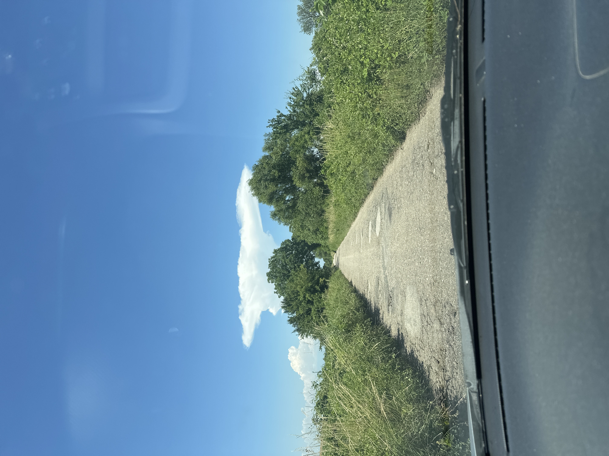 The image shows a view from inside a car driving down a narrow, unpaved road in a rural area. The road is flanked by tall green grass and trees. The sky is blue with a few scattered clouds, one of which is shaped like a mushroom cloud. The dashboard of the car is visible in the foreground. There are no people or animals in the image. The location is a rural road in the vicinity of Muldava, Bulgaria.
