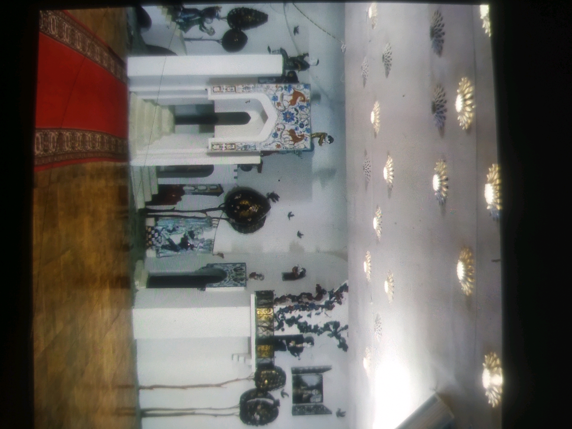 The image depicts the interior of a building, possibly a mosque or a cultural center, in the city of Birlik, Uzbekistan. The scene is dominated by white walls adorned with decorative elements. A red carpet with a patterned border runs along the left side of the frame. In the center, a white architectural structure with ornate blue and gold detailing is visible. Various decorative objects, including what appear to be hanging lamps or chandeliers, are scattered throughout the space. The ceiling is illuminated by a series of decorative light fixtures that resemble sunbursts or floral motifs. The overall impression is one of a richly decorated and possibly historical interior space.
