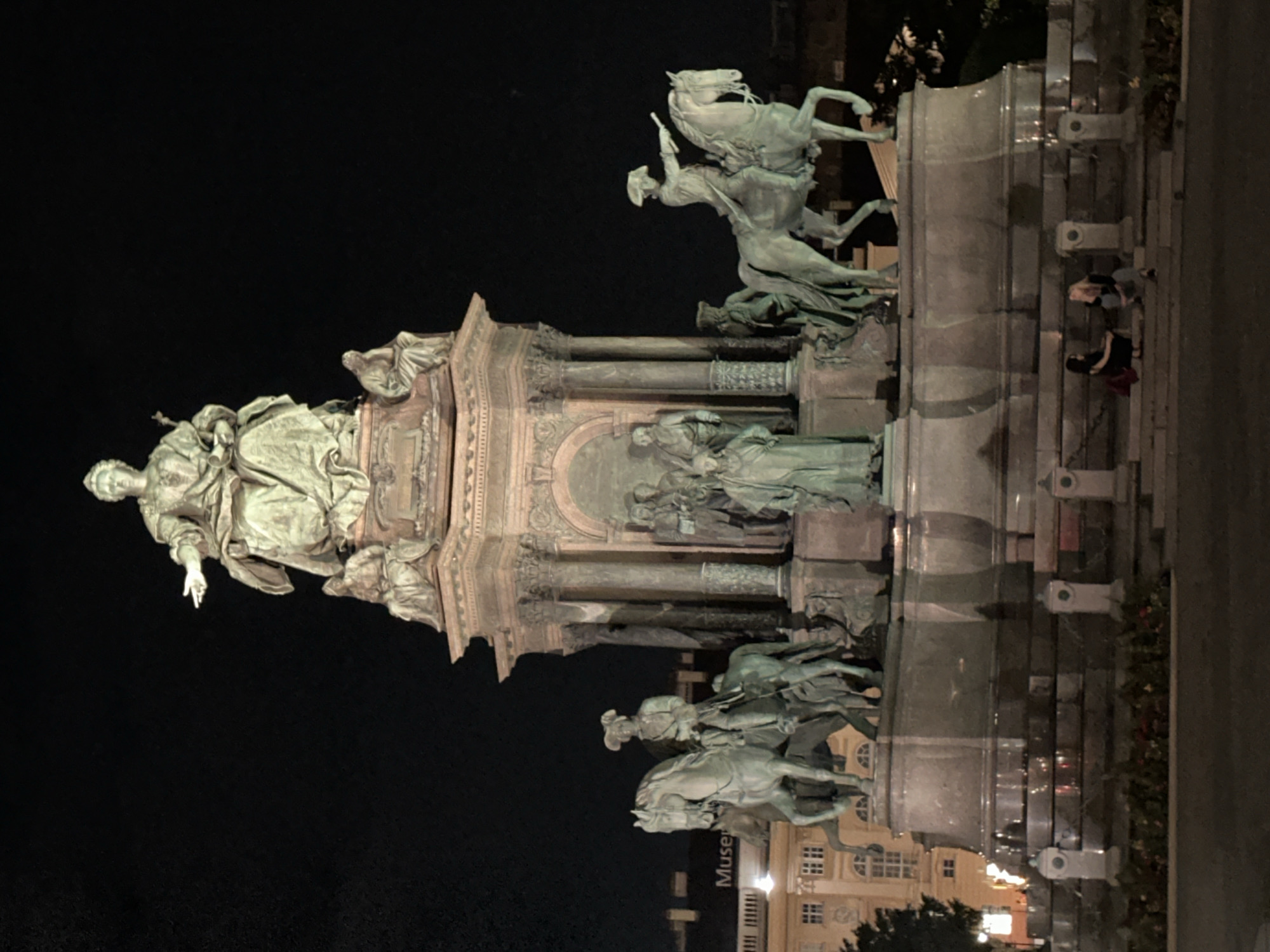 The image depicts the monument to Empress Maria Theresa in Vienna, Austria, at night. The central monument features a large bronze statue of Empress Maria Theresa seated on a throne, with smaller statues and equestrian figures at its base. The monument is illuminated, casting shadows and highlighting the intricate details of the sculptures. In the background, parts of buildings and trees are visible against the dark night sky. In the foreground, two women are seated on the steps of the monument's base, seemingly engaged in conversation. The overall scene conveys a sense of grandeur and historical significance, set against the backdrop of a quiet evening in the city.