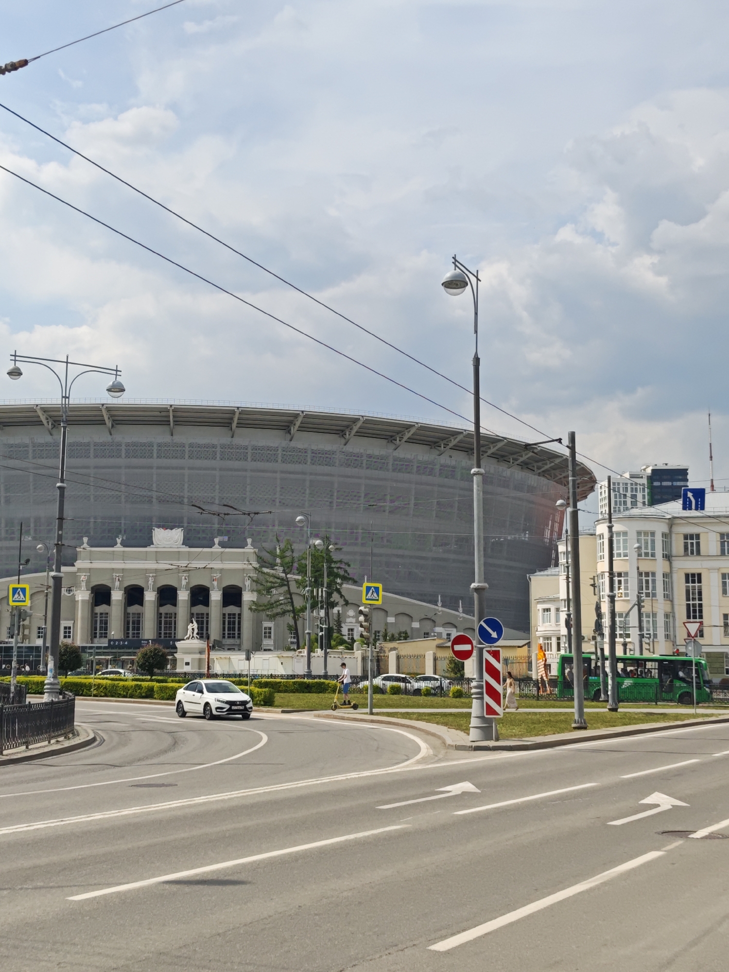The image shows a street scene in Yekaterinburg, Russia. In the background is the Central Stadium, covered in a gray scrim. In the foreground is a road with directional arrows painted on the asphalt. A white car is driving on the road. There are sidewalks on both sides of the road with people walking on them. Several streetlights are present. Traffic signs, including a "No Entry" sign and a directional sign, are visible on a post. A green public transportation vehicle is also visible. The sky is partly cloudy.