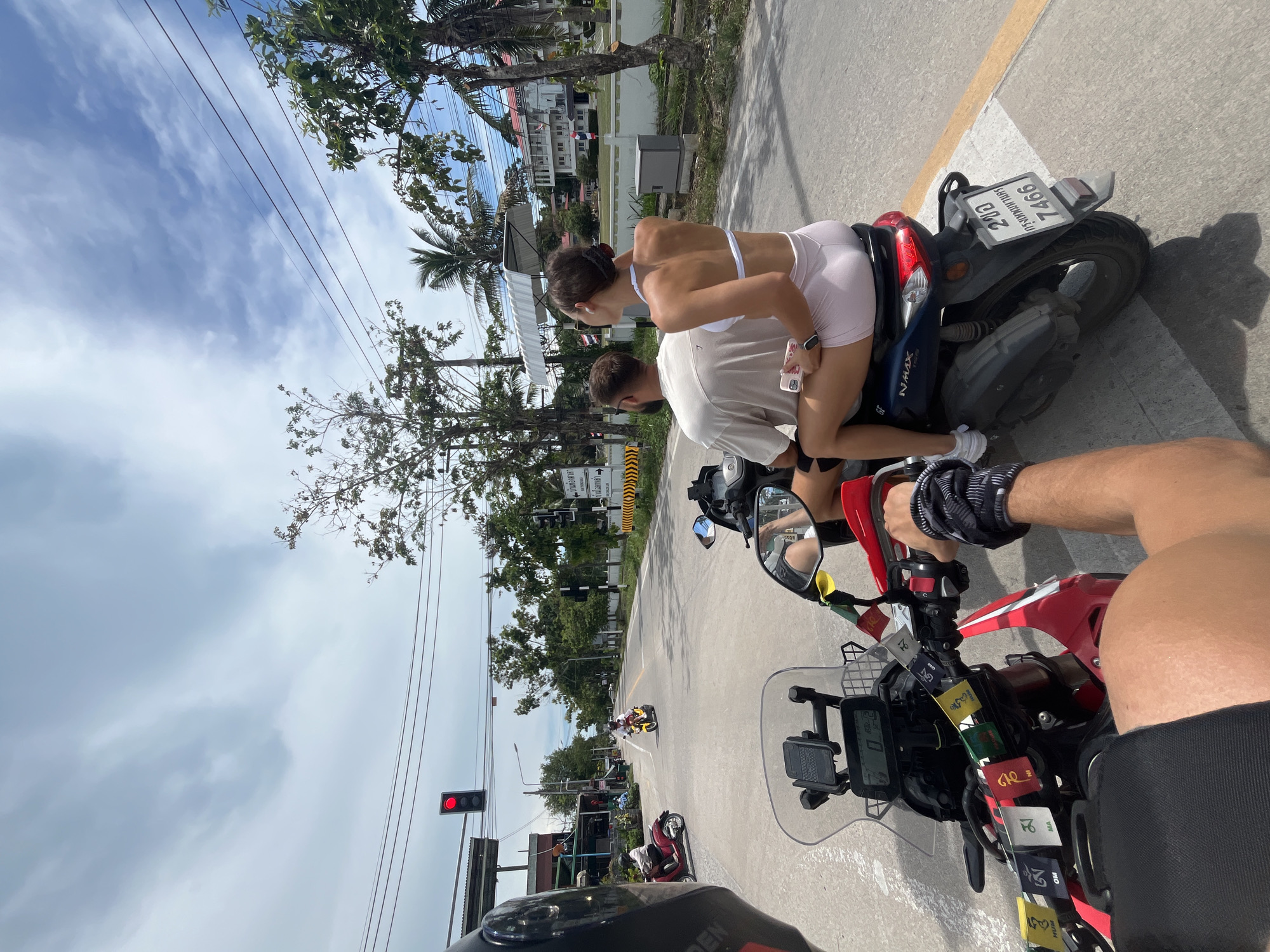 An overhead, wide shot captures a couple riding a scooter on a street in Koh Phangan, Thailand. The scooter is traveling on a paved road with yellow and white lane markings. The man is driving, while the woman sits behind him. Both are dressed in casual clothing, with the woman wearing a white sports bra and pink shorts, and the man in a tan t-shirt and black shorts. A red traffic light is visible in the distance, indicating a potential stop. The background shows buildings, trees, and a blue sky with scattered clouds. The scene appears to be a sunny day, typical of a tropical location.