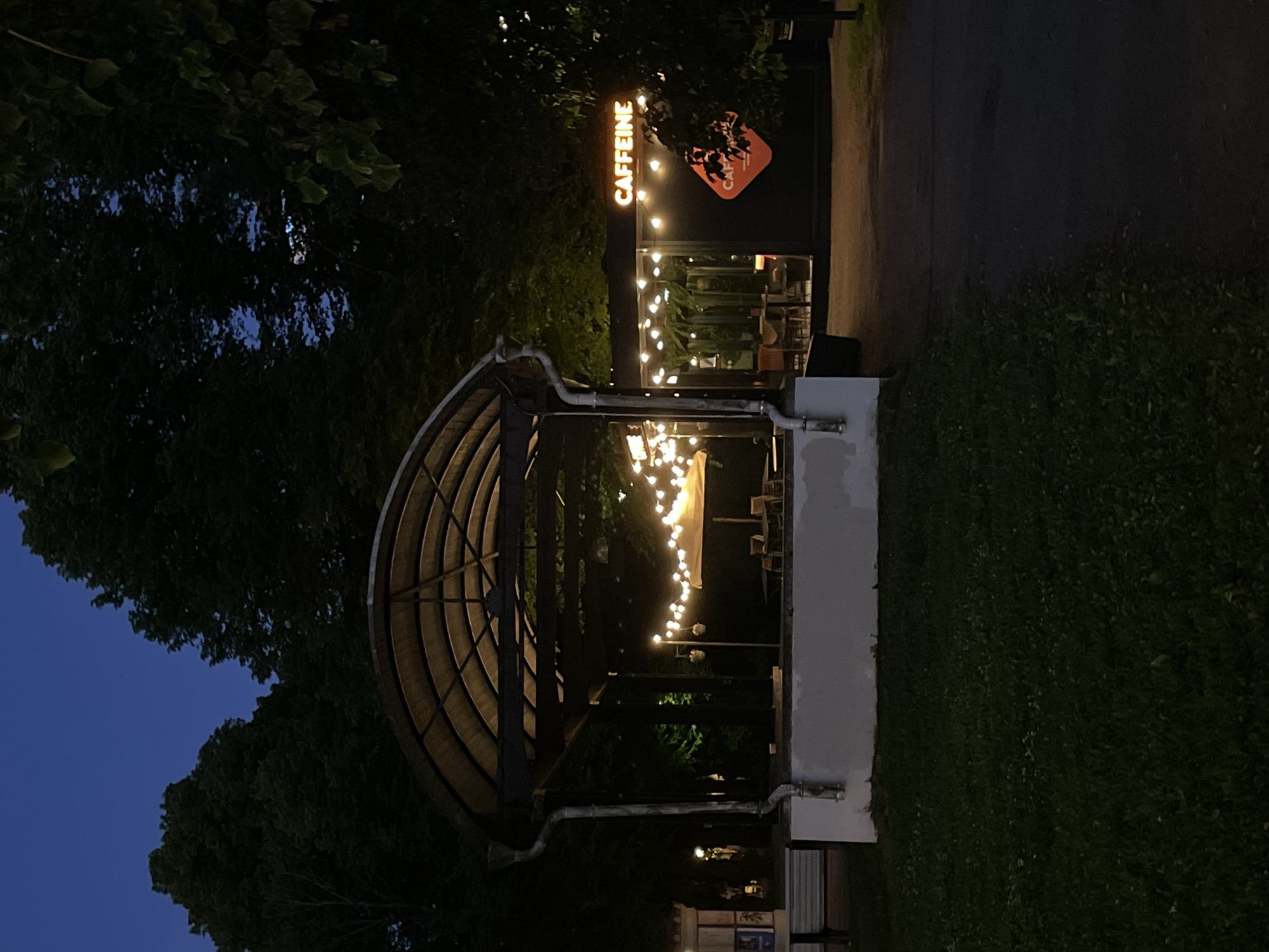 The image depicts an outdoor cafe at dusk in Tallinn, Estonia. A modern, dark-colored cafe building with large windows is visible on the right side of the frame. It is illuminated by strings of warm, white fairy lights draped across its exterior and interior. The cafe's signage, "CAFFEINE," is prominently displayed in orange neon letters above the entrance. A distinctive orange diamond-shaped logo with stylized lettering is also visible on the building's facade.
In the foreground to the left, a covered outdoor seating area with a curved, translucent roof is illuminated by similar fairy lights. Several tables and chairs are set up beneath this canopy. A white, rectangular structure with a gutter pipe running down its side is situated in front of this seating area.
The background consists of lush green trees, their foliage dark against the twilight sky, which has a deep blue hue. The ground is covered by grass in the foreground and a paved pathway or road on the right. The overall atmosphere is peaceful and inviting, suggesting a popular spot for evening relaxation or socializing. No people are visible in the image.