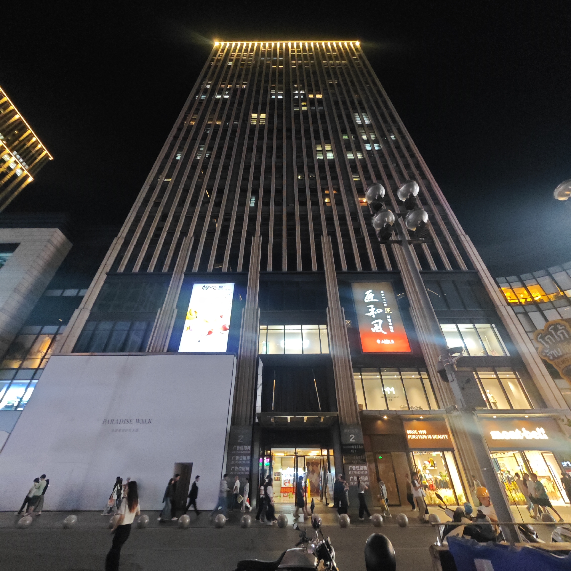 The image captures a nighttime street scene in what appears to be the Yuzhong District of China, specifically a commercial area in Chongqing. A tall building with a distinctive facade featuring vertical structural lines dominates the view, its top illuminated. Below this building, retail shops and storefronts are visible, with signs like "PARADISE WALK" and brands such as "mont-bell" displayed.
People are seen walking along the sidewalk, some individually and others in groups, indicating pedestrian activity. Various objects, including motorcycles parked near the curb and bollards delineating the pedestrian area, add to the urban environment. The overall atmosphere is one of a bustling commercial district at night, marked by bright lighting and a mix of architectural styles.
