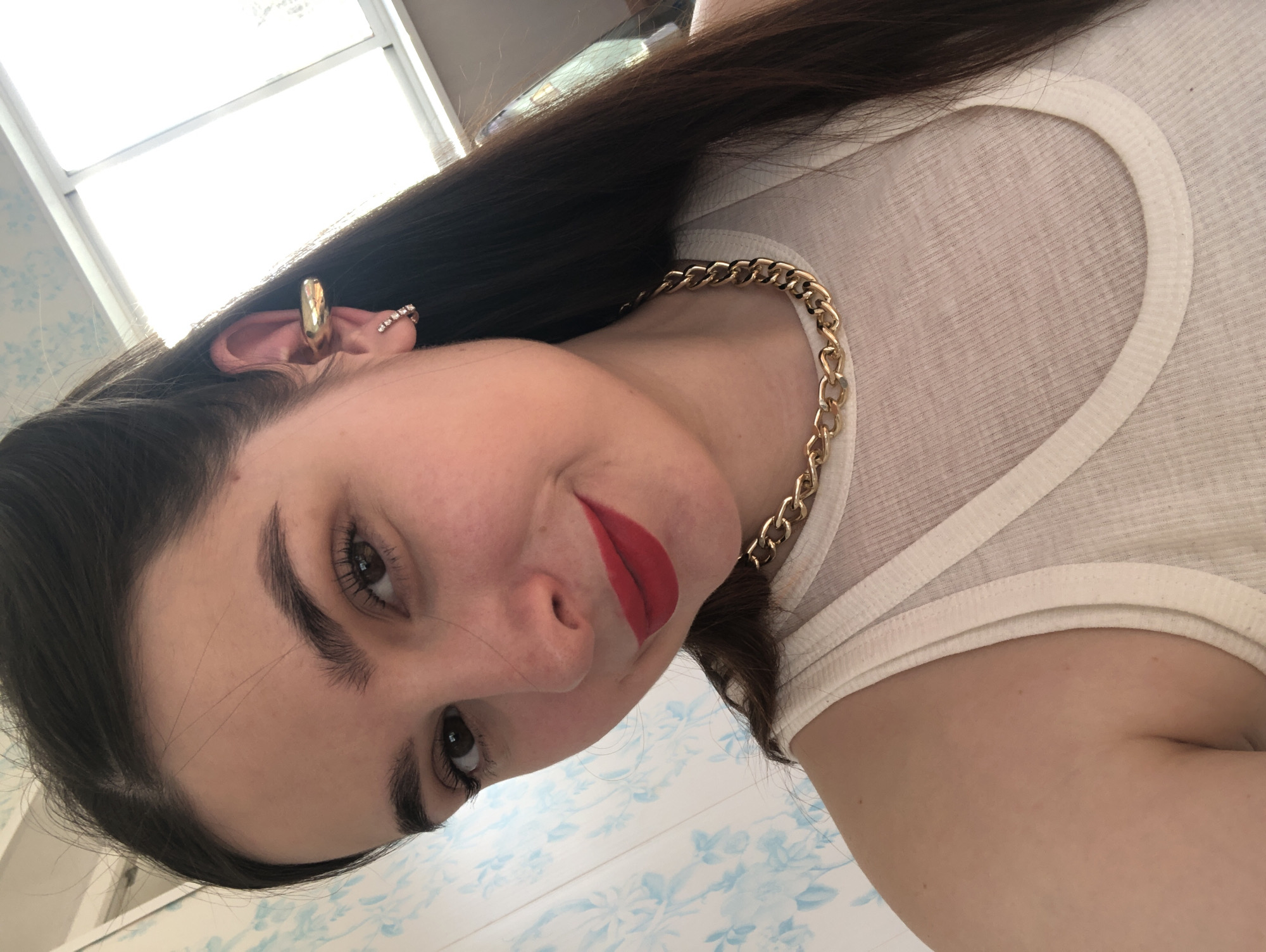 A young woman with long, dark hair is depicted in a close-up, angled portrait, likely a selfie. She has dark eyes, well-groomed eyebrows, and is wearing bright red lipstick. Her expression suggests a subtle, pleasant smile.
She is adorned with jewelry and clothing: a prominent gold-colored chunky chain necklace rests around her neck, and she wears a white ribbed sleeveless top or tank top. On her visible ear, she has two distinct pieces of jewelry: a large, shiny gold-colored ear cuff, and a smaller ear cuff or earring embellished with what appear to be small, clear sparkling stones.
The setting appears to be indoors during daytime, indicated by bright natural light coming from a window visible in the background. A distinctive light blue and white patterned surface, possibly wallpaper or tile, is seen on what looks like a lower wall or floor. There is no visible text in the image. The location context is Noviy, Russia.