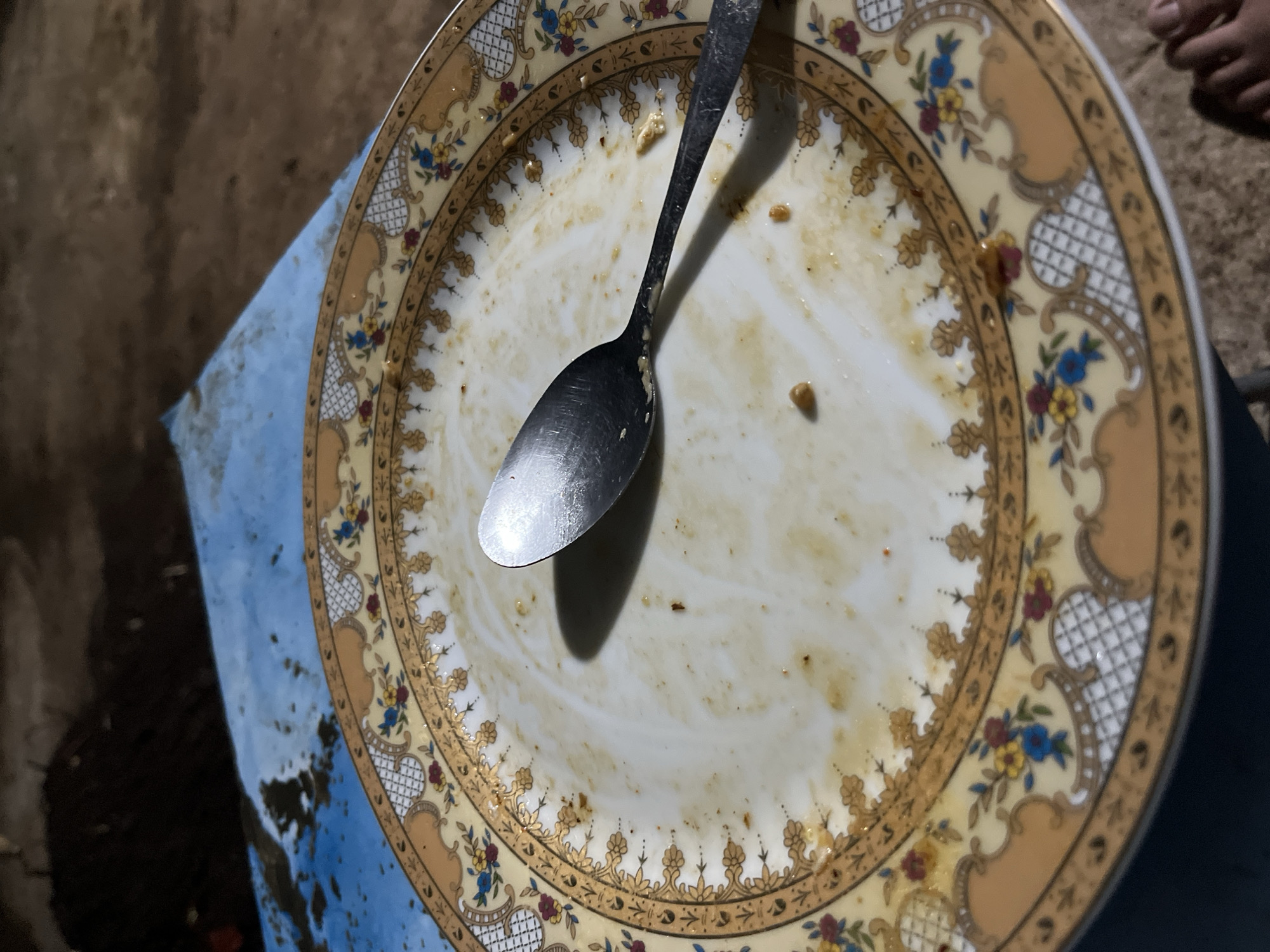 A close-up, overhead shot shows a partially eaten meal on a decorative plate. A silver spoon rests on the plate, casting a shadow. The plate has a floral and gold trim design. The background consists of a blue surface and brown dirt or rough ground. A person's toes are visible at the bottom right of the image. The image appears to be taken in an outdoor setting in Central Jakarta, Indonesia, likely in a modest or informal environment given the unpolished background.