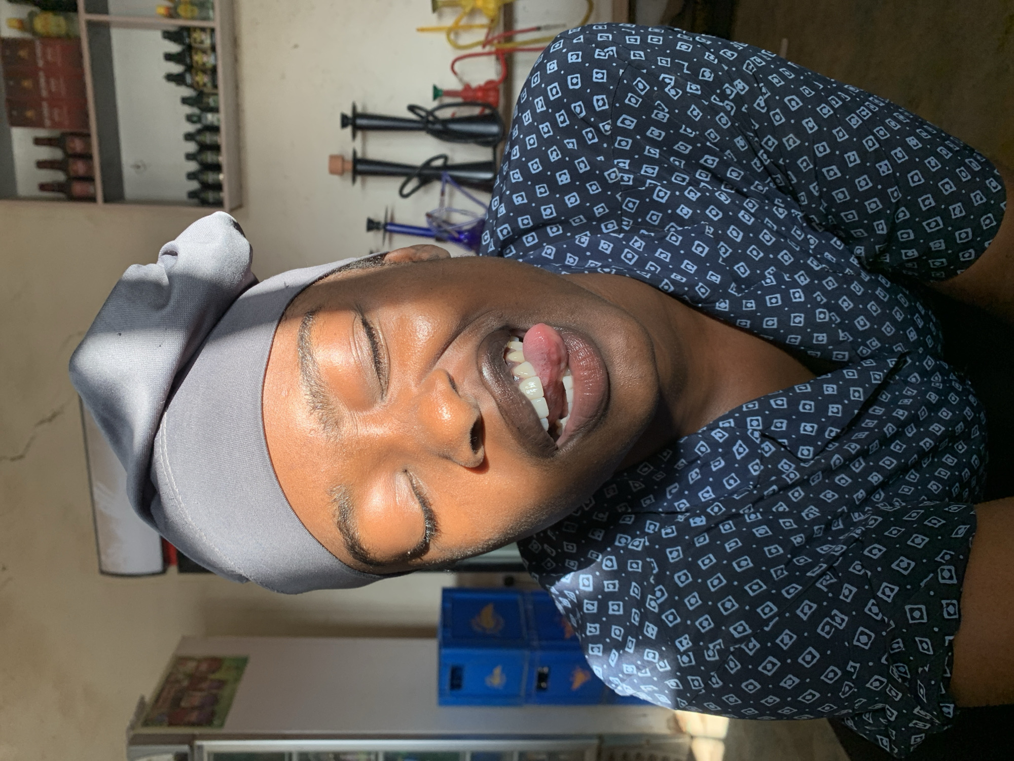 A person wearing a grey head covering and a dark blue shirt with a pattern is sticking out their tongue. In the background there are bottles on a shelf, hookahs, and blue crates. The location is Kabong, Nigeria.