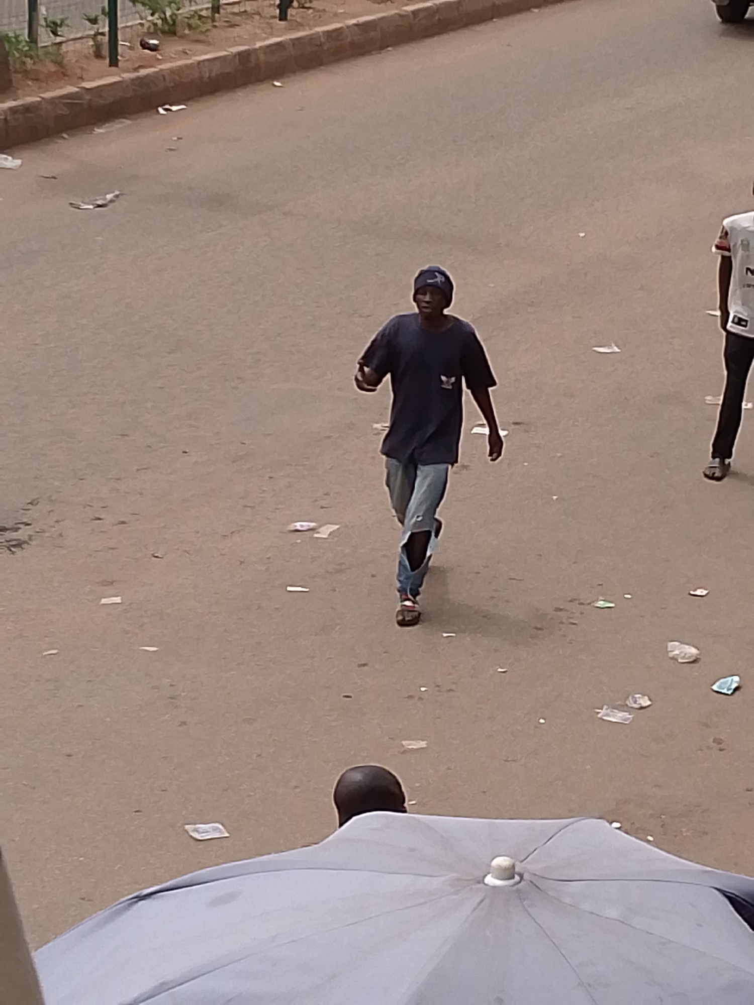 The image shows a street scene with a person wearing a dark blue shirt, ripped jeans, and a beanie walking towards the camera. Another person wearing a white shirt is partially visible to the right. The street appears to be littered with paper and other debris. A gray umbrella is visible in the foreground. The location appears to be a city street in Unknown, Unknown.