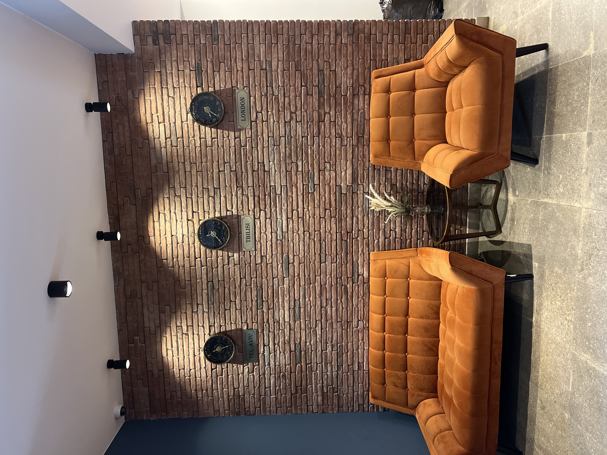 The image shows a waiting area with two modern, orange velvet upholstered chairs and a matching loveseat against a textured brick wall. Three round clocks, each with a city name plate below, are mounted on the wall: "TEL-AVIV", "TBILISI", and "LONDON". A small, round glass-top table with a metal base sits between the loveseat and the chair, holding a decorative arrangement of dried grass. The floor is tiled with large, grey square tiles. Several black cylinder-shaped lights are mounted on the white ceiling, and a security camera is visible on the ceiling to the left. The overall aesthetic suggests a contemporary, perhaps boutique, hotel lobby or reception area. The presence of the "TBILISI" clock strongly indicates this scene is located in Tbilisi, Georgia.