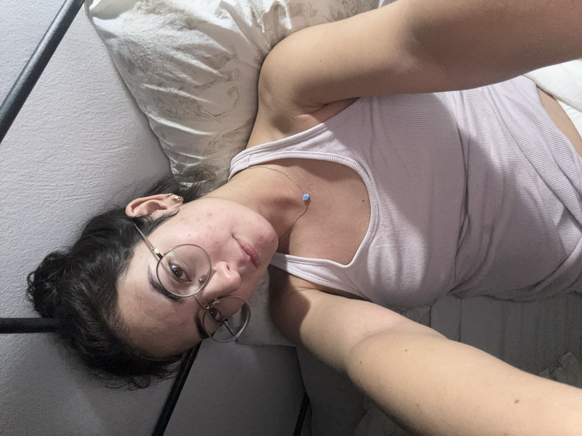 A woman lies on her side in bed, wearing a light pink tank top and round eyeglasses. She has dark hair pulled back in a bun and a small necklace with a blue pendant. The background includes a white pillow and a gray wall with a metal bed frame. The scene is indoors and no information is provided to suggest it is Puebla, Mexico.