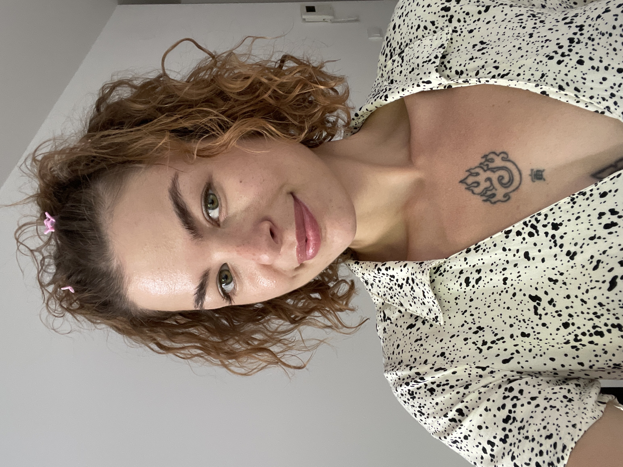 A young, light-skinned woman with curly reddish-brown hair is pictured in a close-up, slightly angled view. She has green eyes and is wearing a white dress with a black speckled pattern. A small tattoo is visible on her upper chest. Her hair is styled with a couple of pink clips near her forehead. The background is a plain white wall. There is no indication of the city of Boreti, Montenegro.
