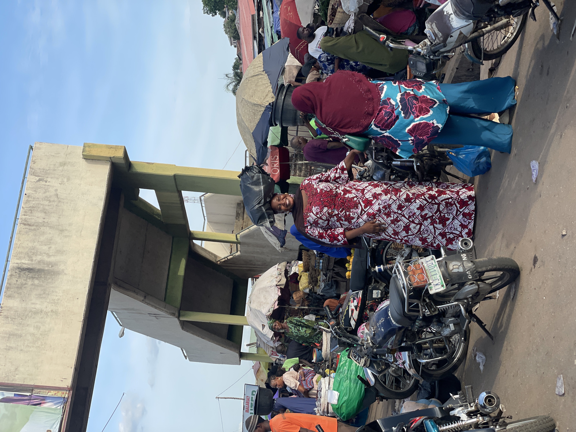 The image captures a bustling scene in a marketplace in Lokoja, Nigeria. A woman in a red and white patterned dress stands facing the camera, with other individuals in the background partially obscured by umbrellas and market stalls. Several motorcycles are parked closely together on the street. Overhead, there is a concrete pedestrian walkway. The overall atmosphere is busy and crowded, characteristic of a marketplace environment.