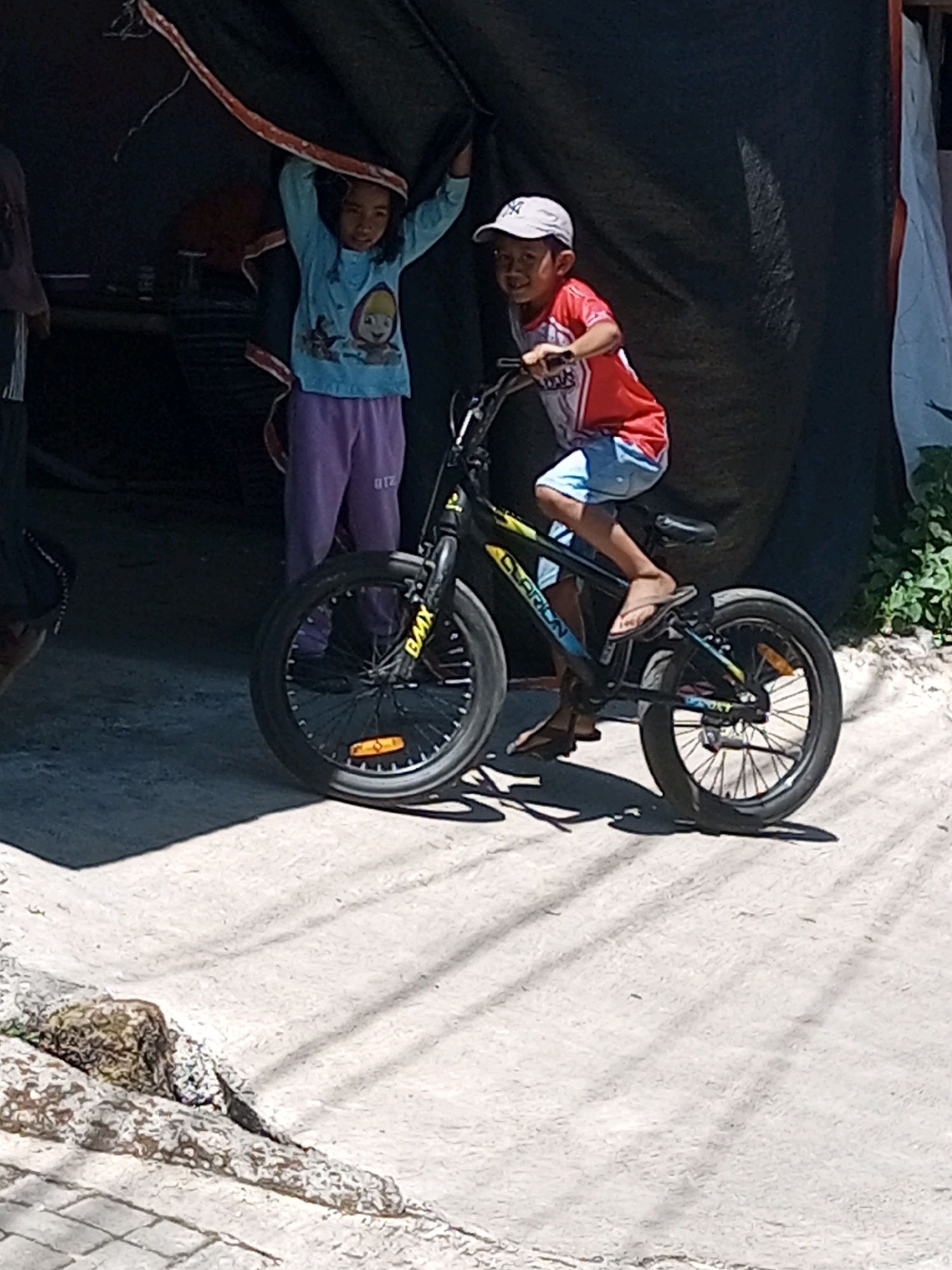 Two children are near a bicycle in a sandy area, with a dark tarp covering part of the background. The child on the left, a girl, stands and holds a draped orange and brown fabric above her head. She is wearing a light blue long-sleeved shirt with cartoon characters and purple pants. The child on the right, a boy, is seated on a black BMX-style bicycle, facing the viewer. He is wearing a red and white t-shirt, blue shorts, a white baseball cap, and sandals. The bicycle has yellow and blue accents on its frame. The ground is a mix of sand and some paved elements in the foreground, with shadows cast from above. The overall scene appears to be outdoors, possibly in a rural or less developed urban area. This image was taken in Andongsili, Indonesia.